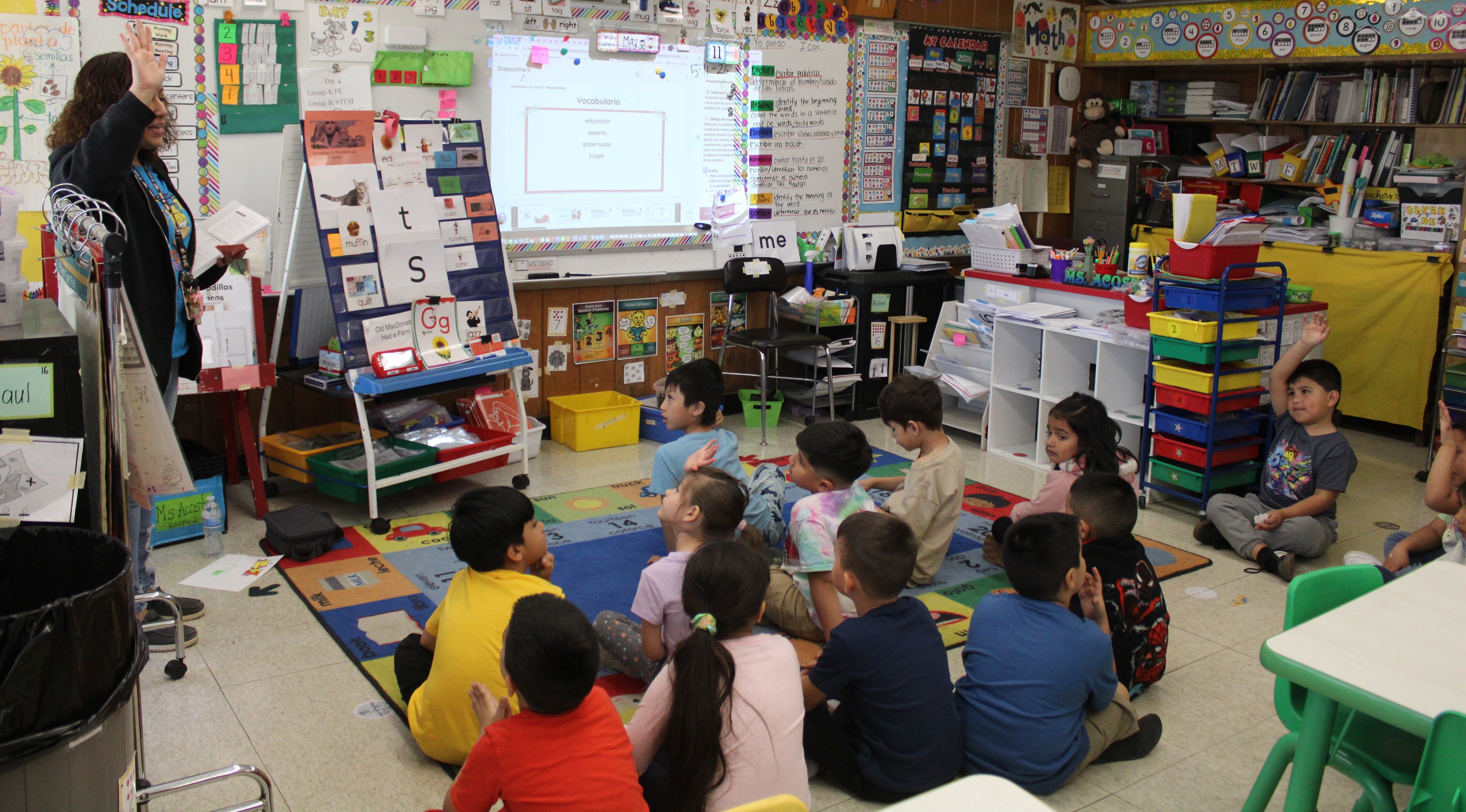 Engaged students sitting on the floor around a teacher, raising hands to answer a question.