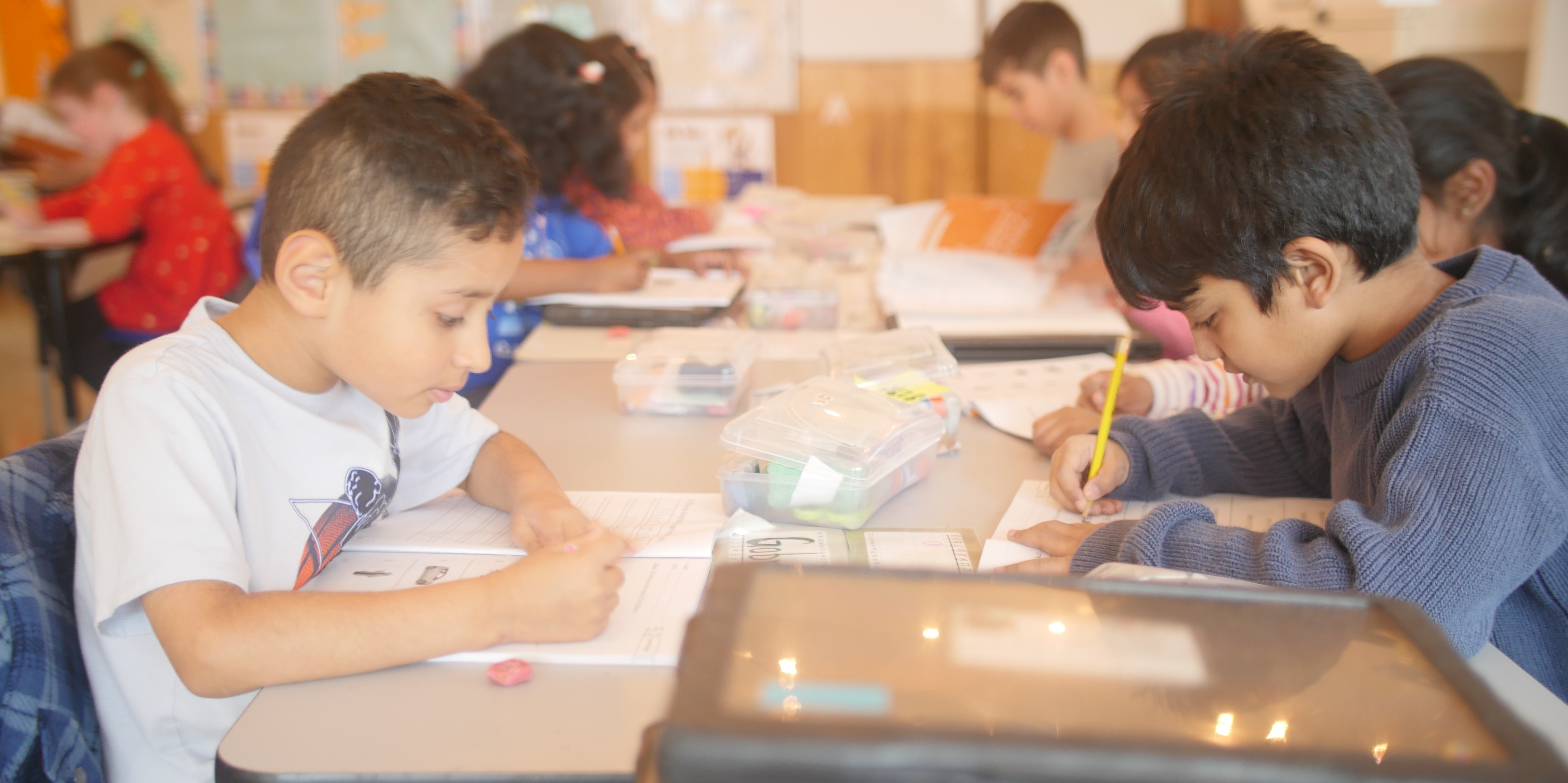 Students sit at grouped tables, holding pencils and focused on working through their classroom worksheets