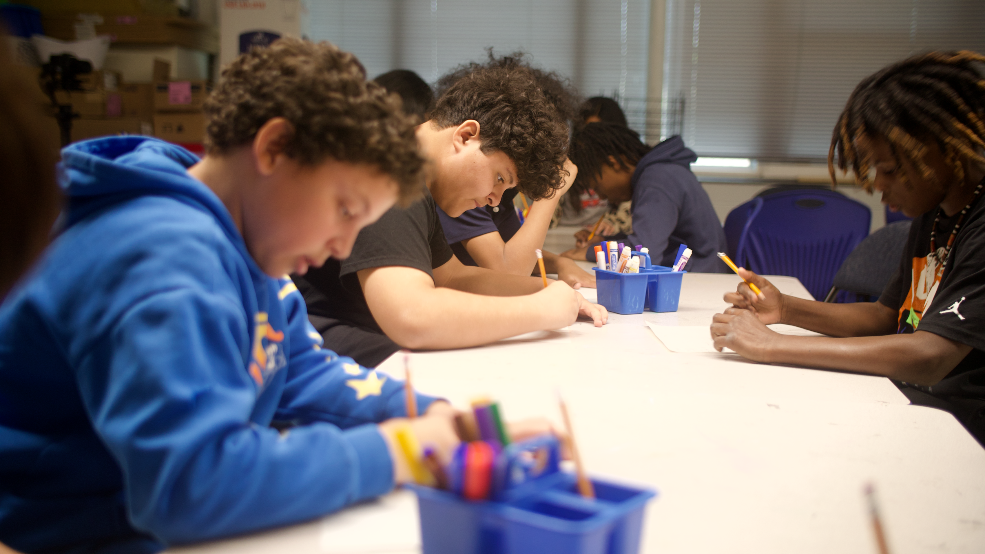 Students sit at grouped tables, holding pencils and focused on working through their classroom worksheets
