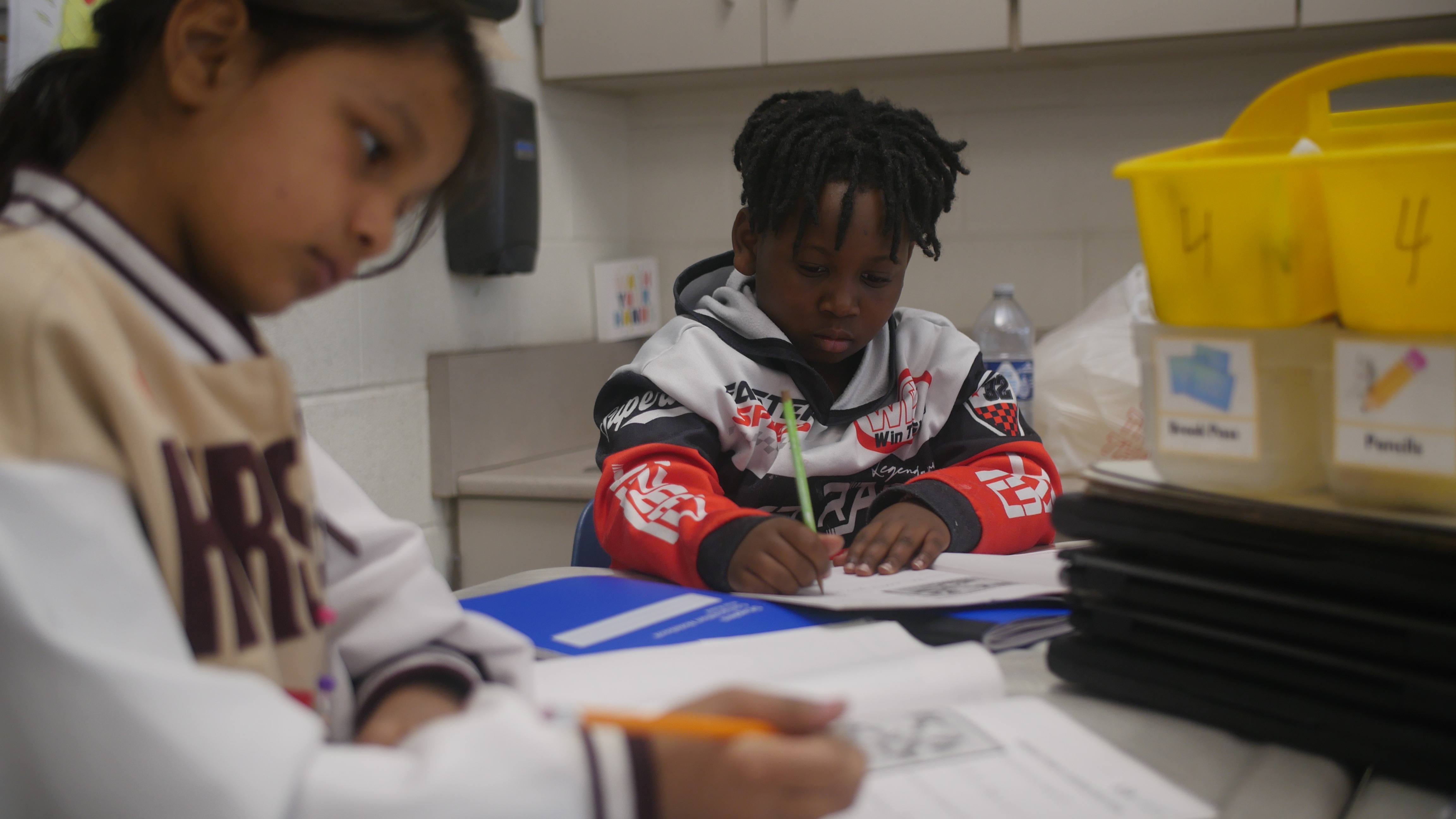 Two students sit at tables, holding pencils and focused on working through their classroom worksheets