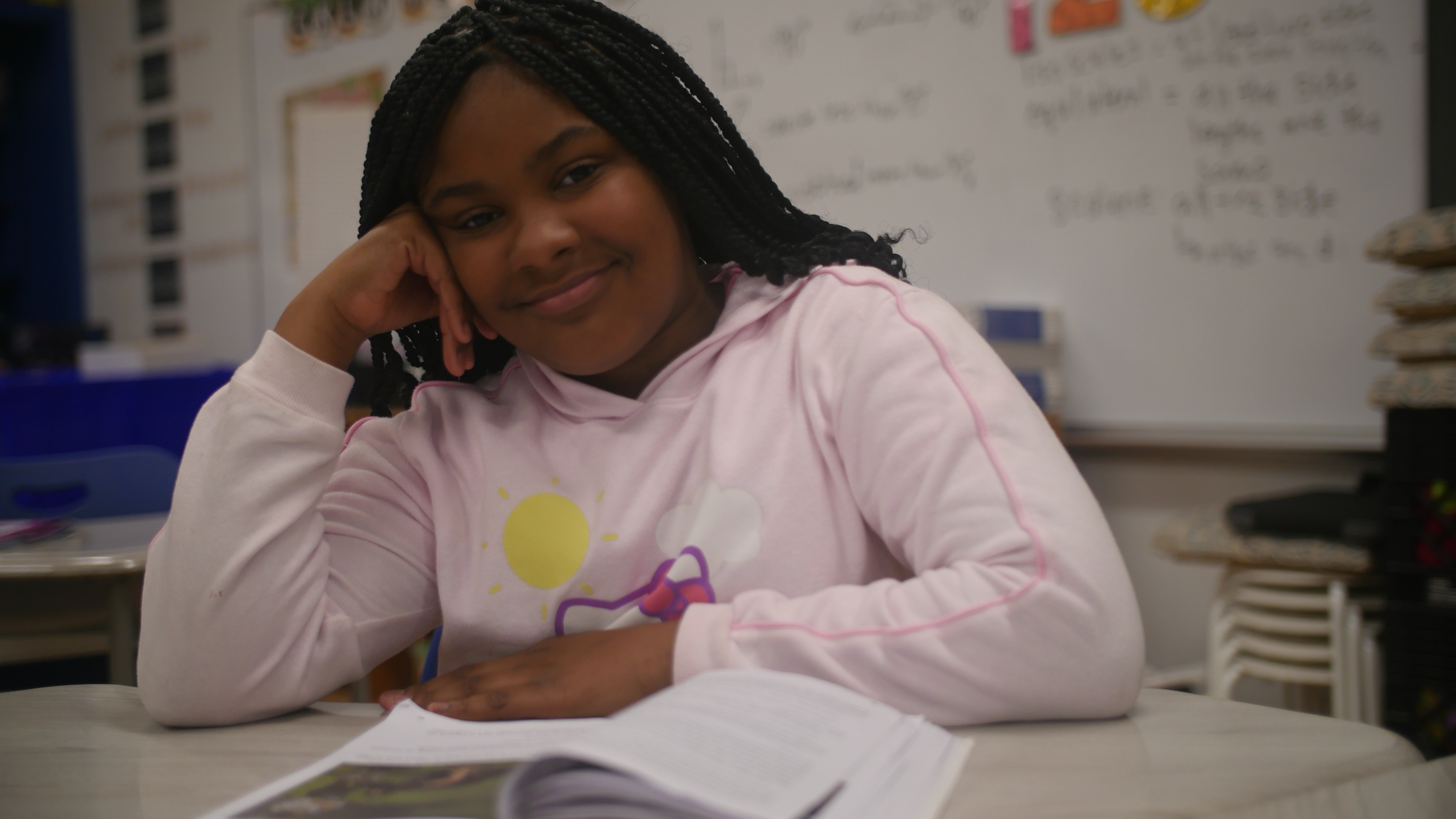 A smiling student sits at her desk with an open book, ready to learn.