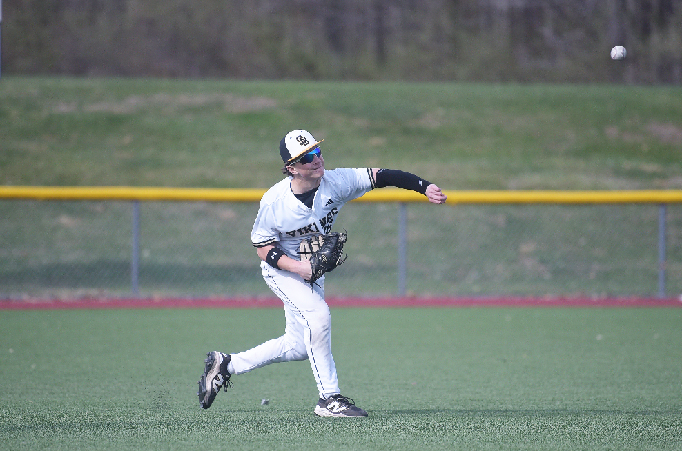 Player in a baseball uniform catches a ball mid-air on a field with a fence and grassy area.