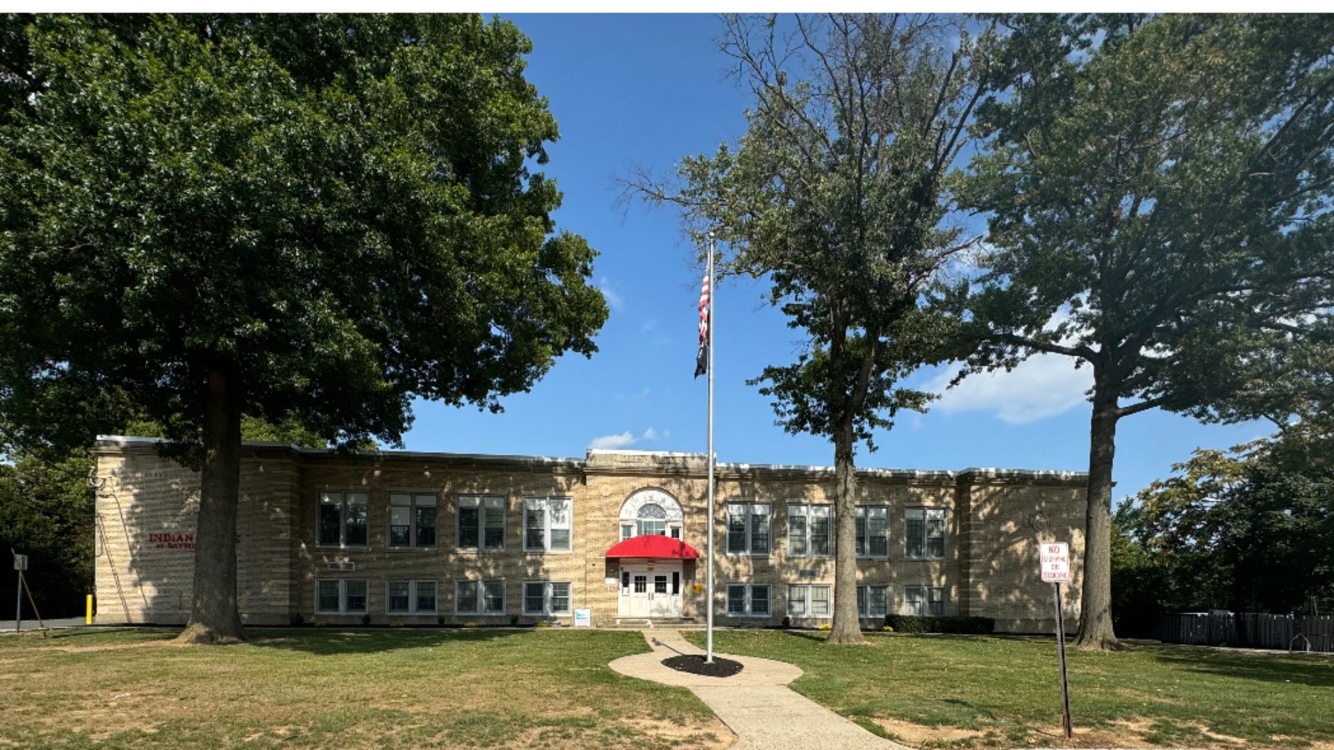 Large building with a red awning over the entrance. Flagpole with flag beside a walkway. Trees with green leaves surround the building.