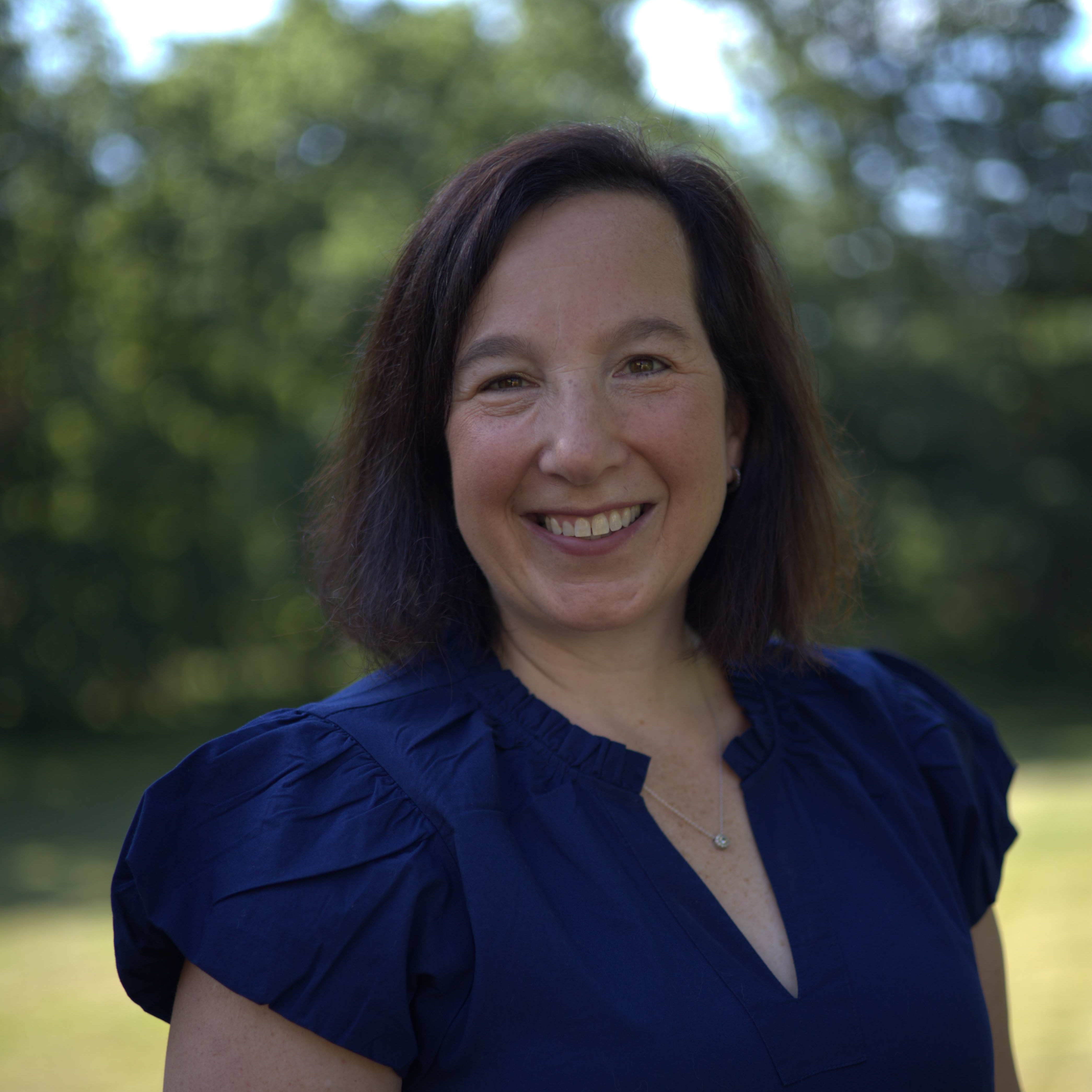 A woman with short hair wearing a navy blue blouse stands outside on a porch with greenery behind her.
