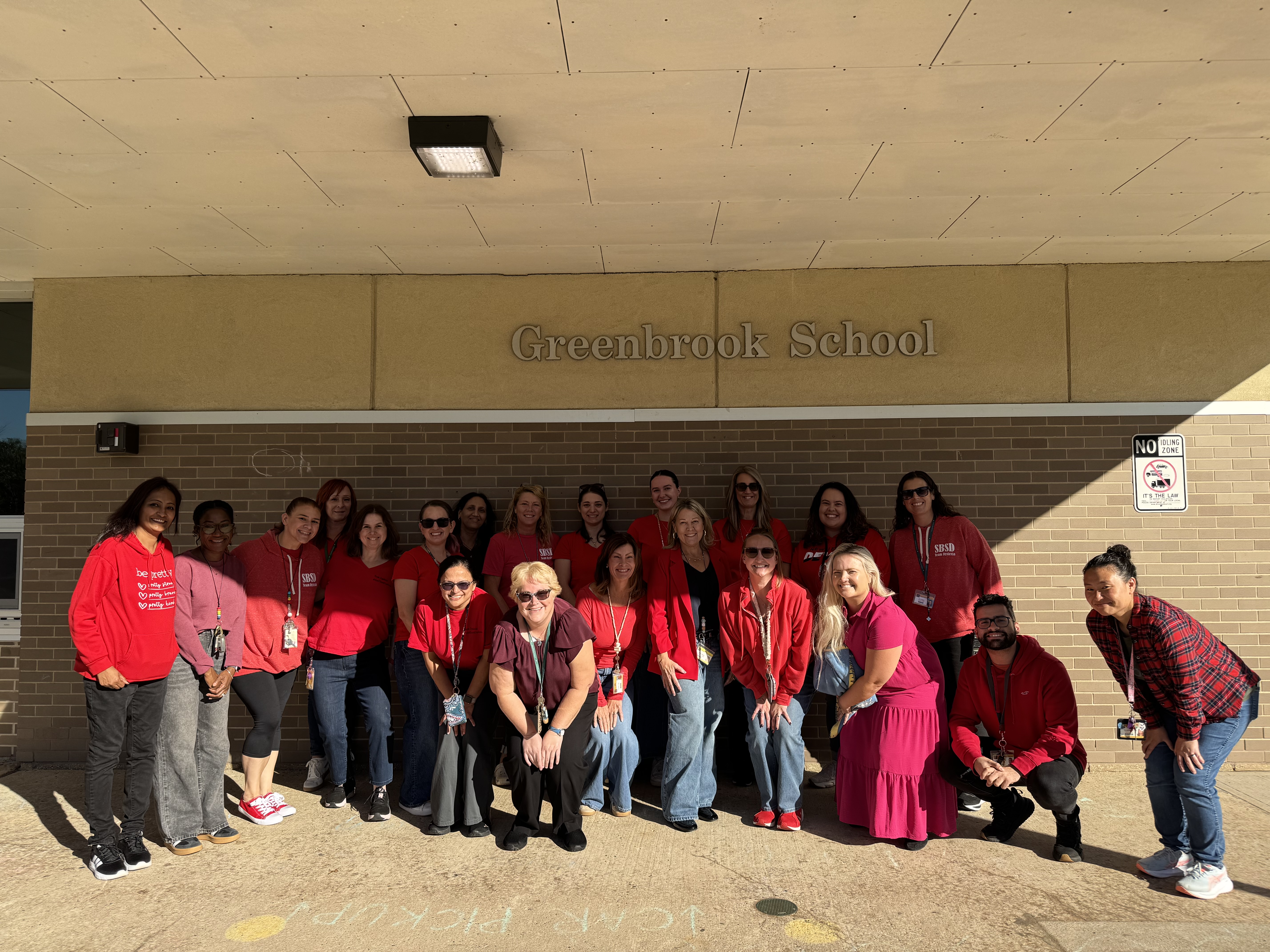 Group of people wearing red tops standing outside near a building with a sign that reads "Greenbrook School"