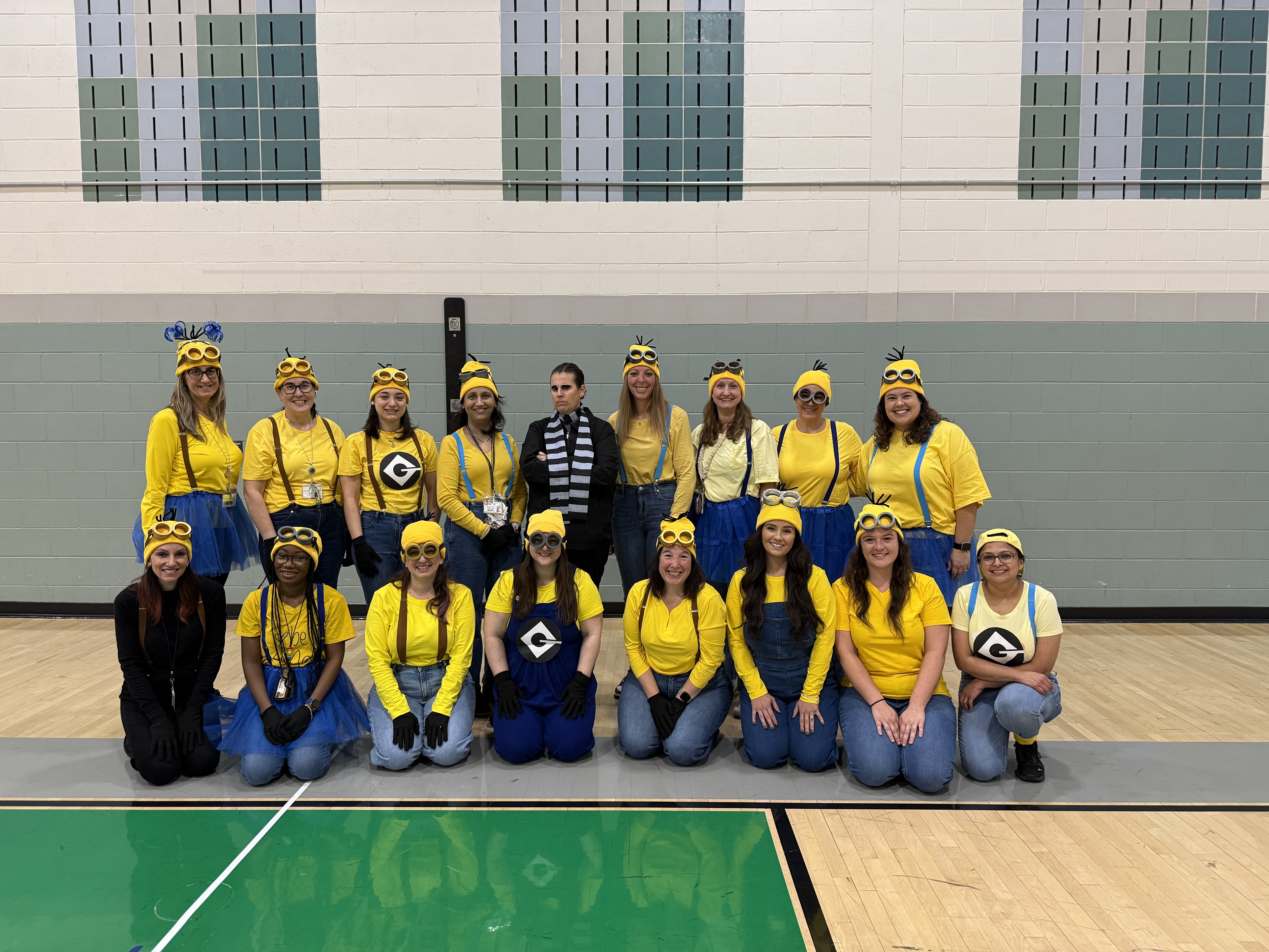 People dressed in yellow and black costumes with eyeless heads pose in a gymnasium with wood floors.
