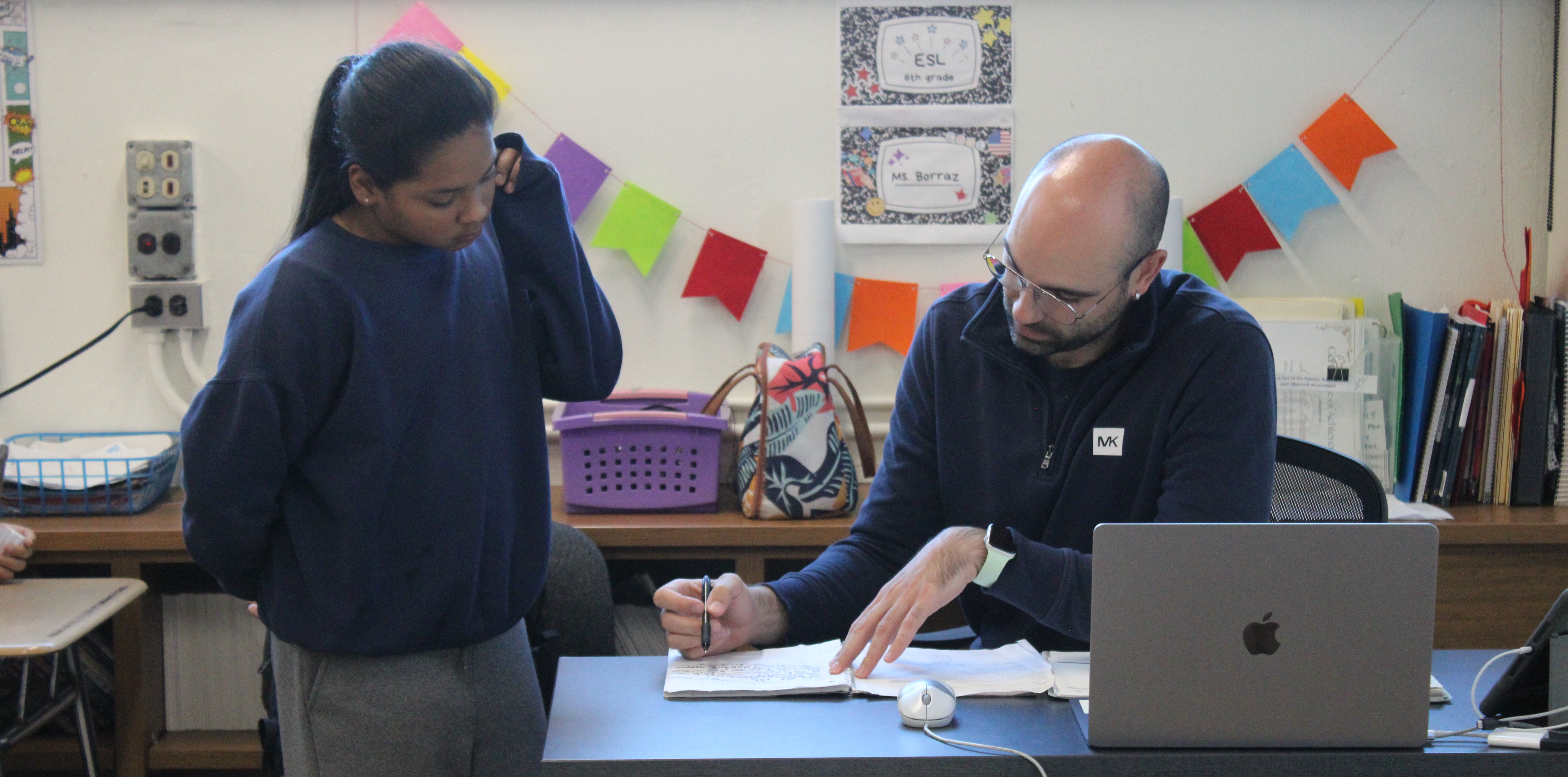 Teacher at a desk reviewing a student's notebook assignment.