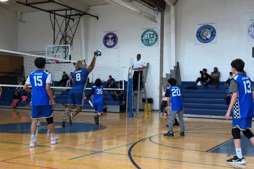 A school volleyball team competes in a match on an indoor court, with the school mascot dressed in costume on the court playing alongside the team