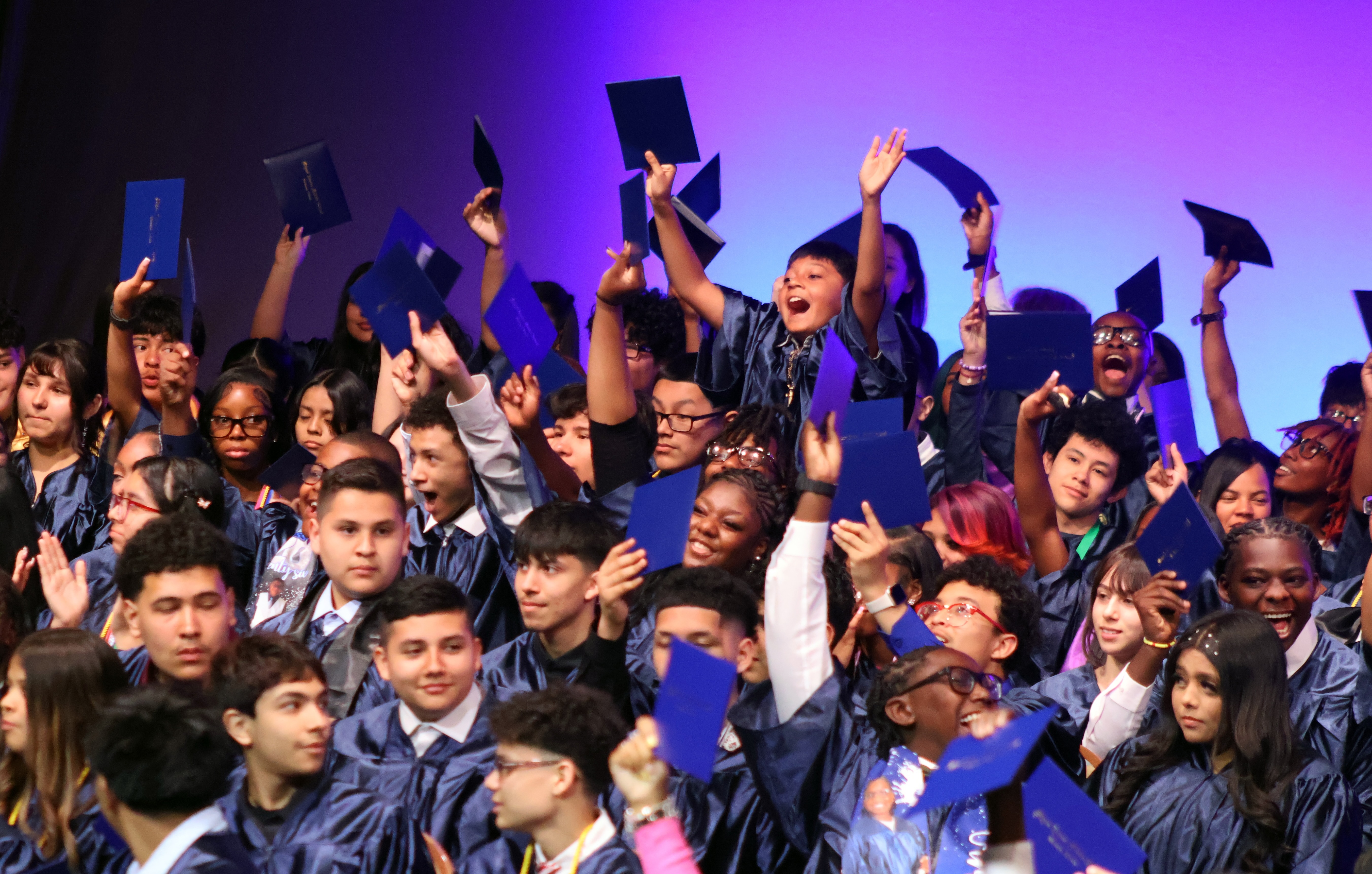 A group of middle school graduates joyfully celebrate their graduation, raising their hands in the air while holding up their diplomas