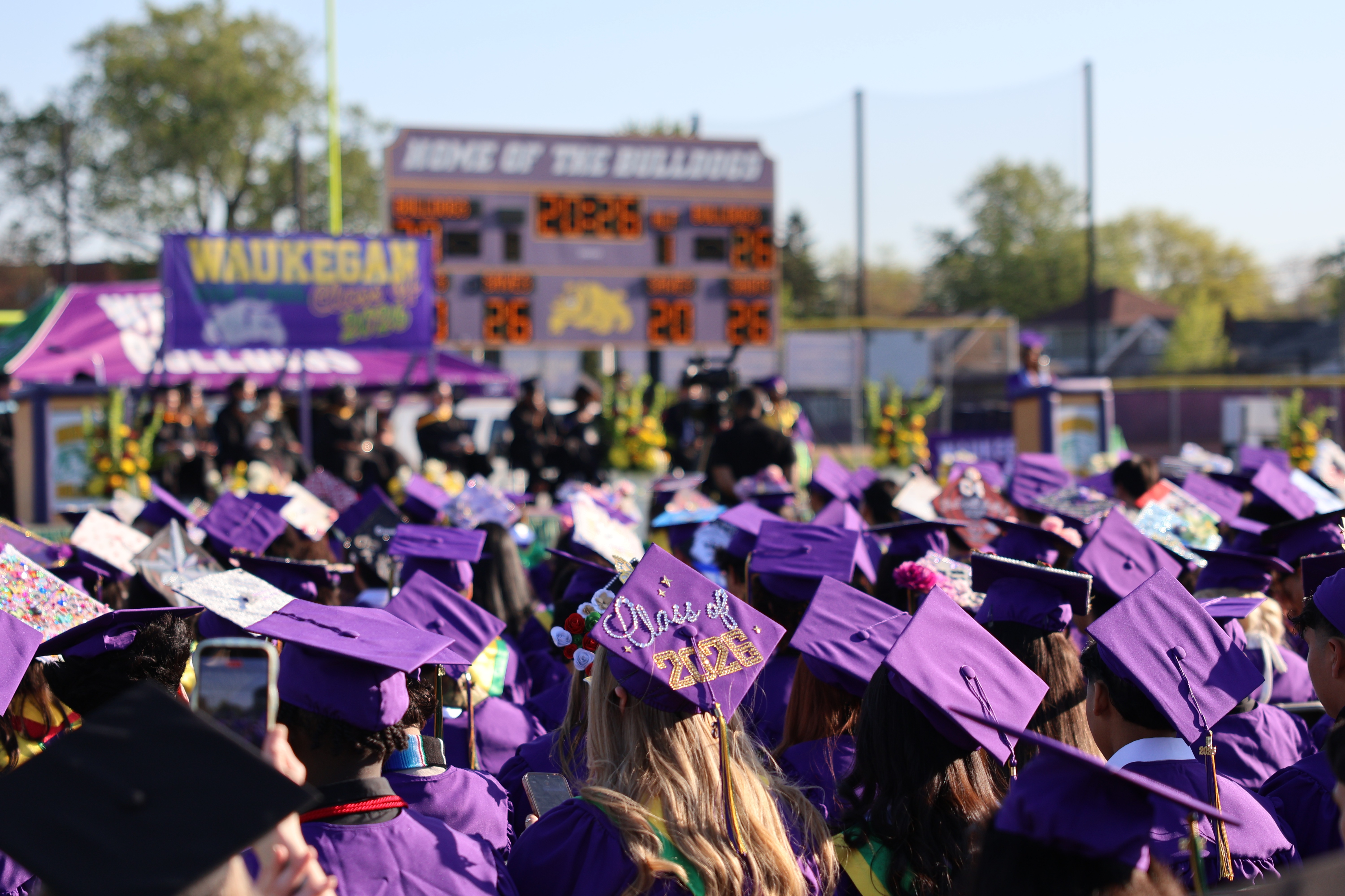 Graduation ceremony view from behind the graduates, showing the backs of their decorated caps with the stadium scoreboard displaying '2026' in the background.