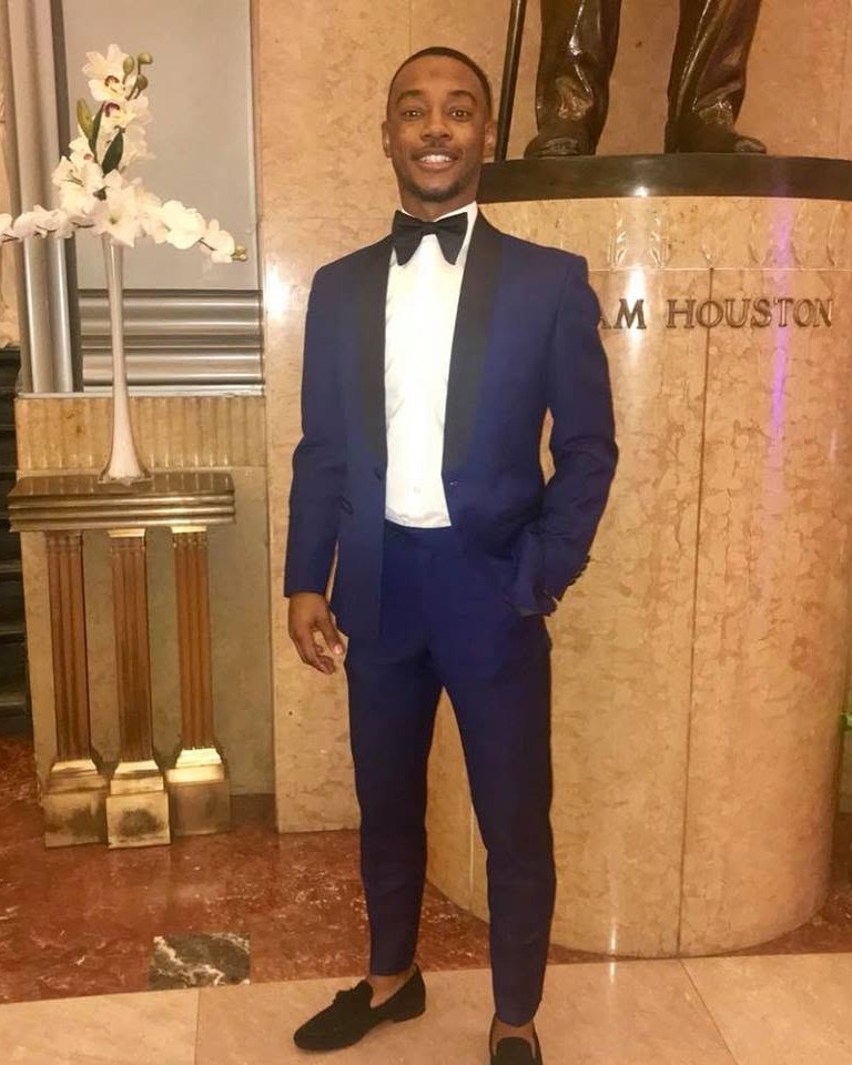 A man in a dark blue tuxedo, white shirt, and black bow tie stands against a marble wall.