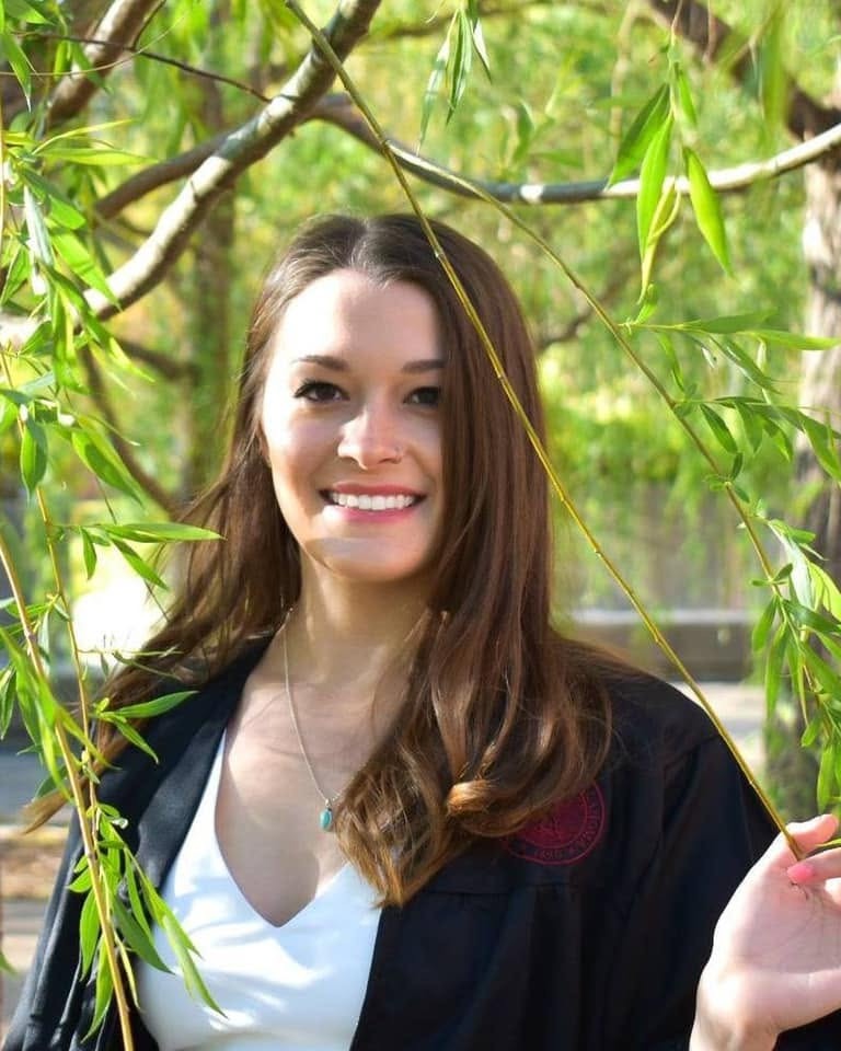 Woman with dark hair, wearing white shirt and black jacket, smiling, standing under a willow tree.
