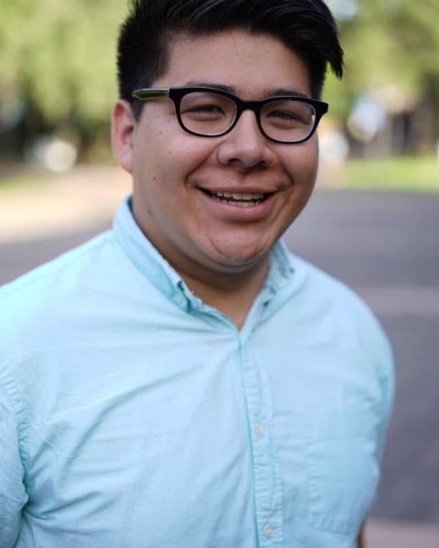A man with glasses and a light blue shirt smiles for the camera. Trees and a pathway are in the background.