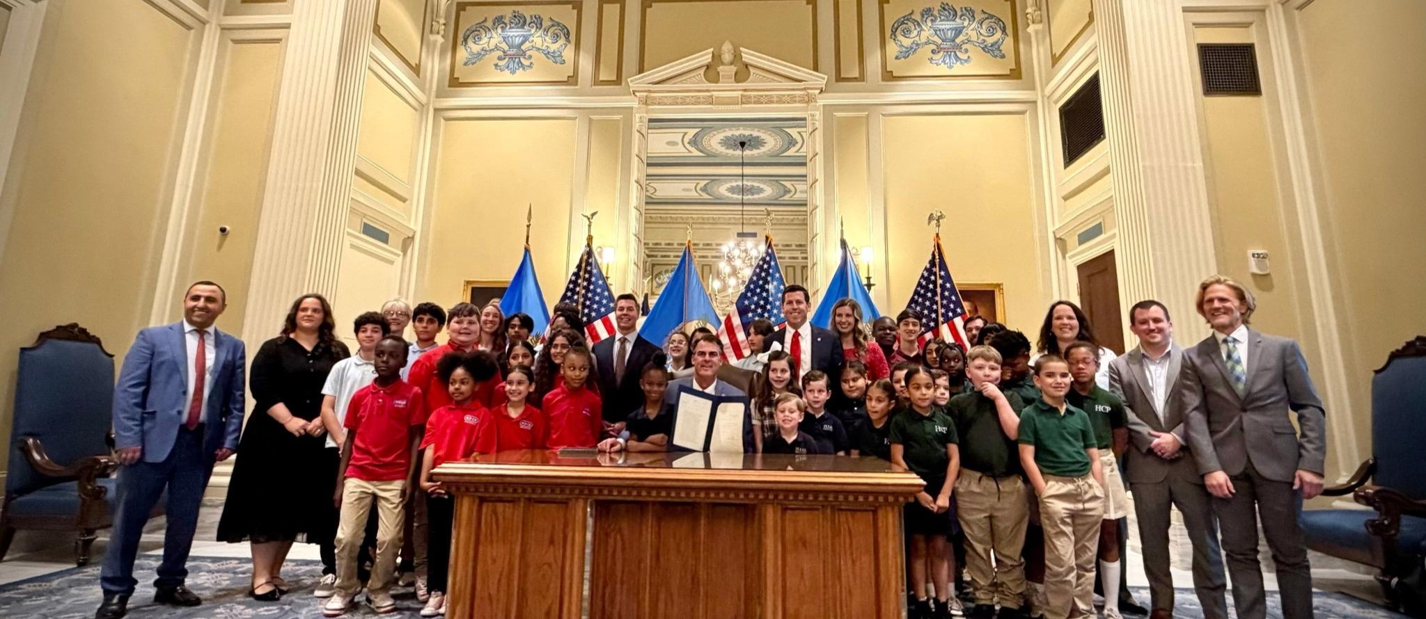 People stand in a conference room with a wooden desk, flags, and formal attire. A group of children in red shirts gather around a person holding a document.