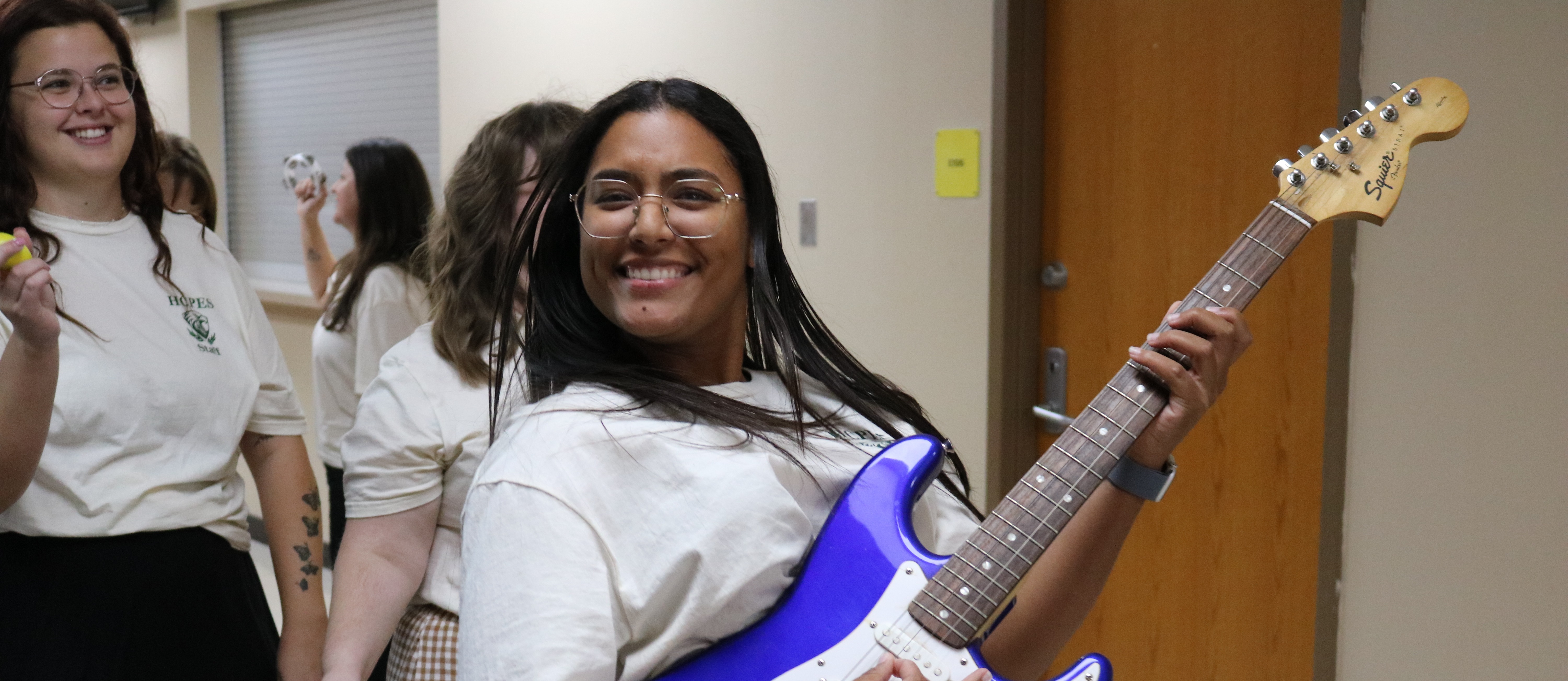 A woman holds a blue and white electric guitar in a hallway. Behind her, three people walk, one holding a camera.