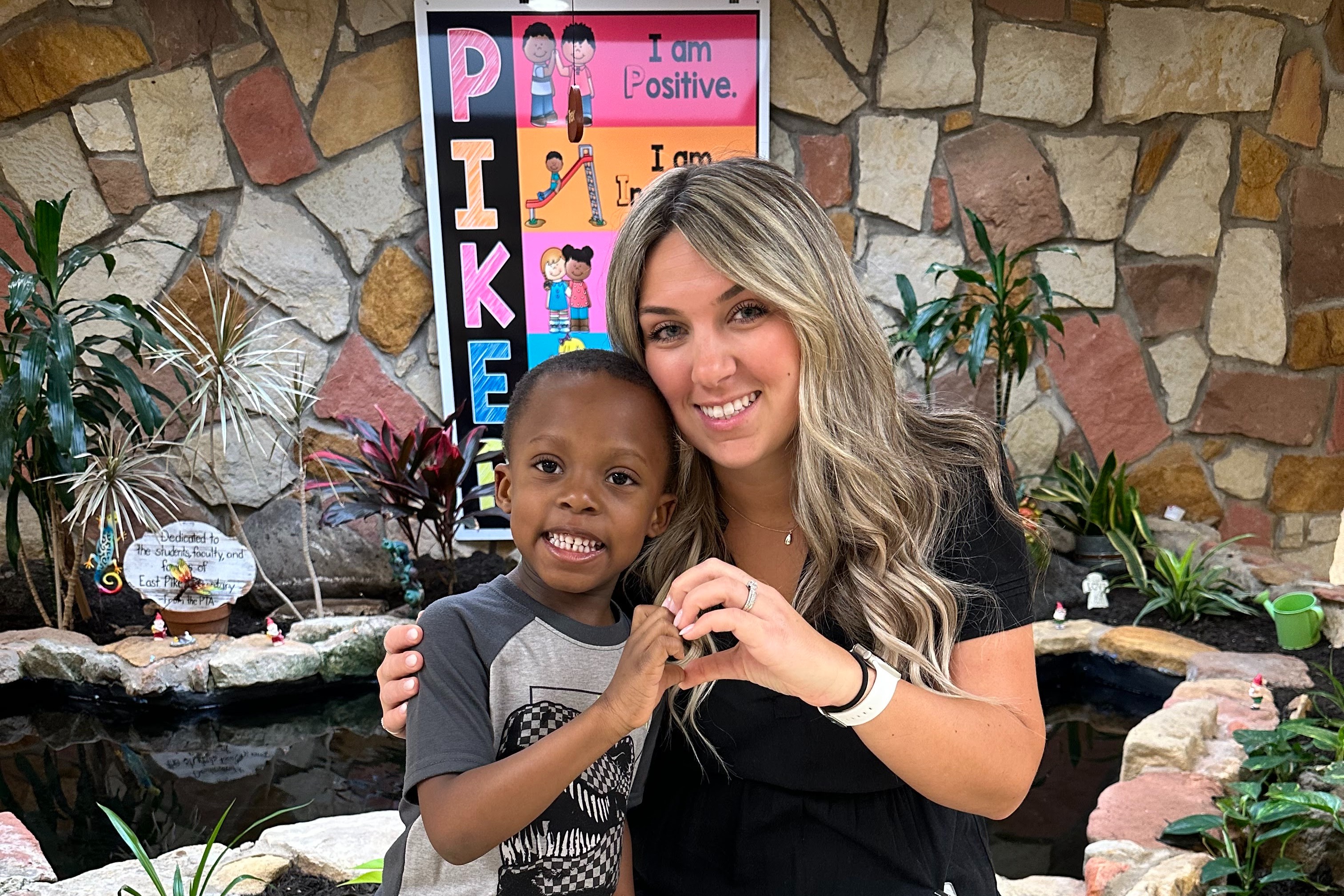 A smiling woman in a black dress poses with a child wearing jeans and sneakers. They stand in front of a stone wall with plants.