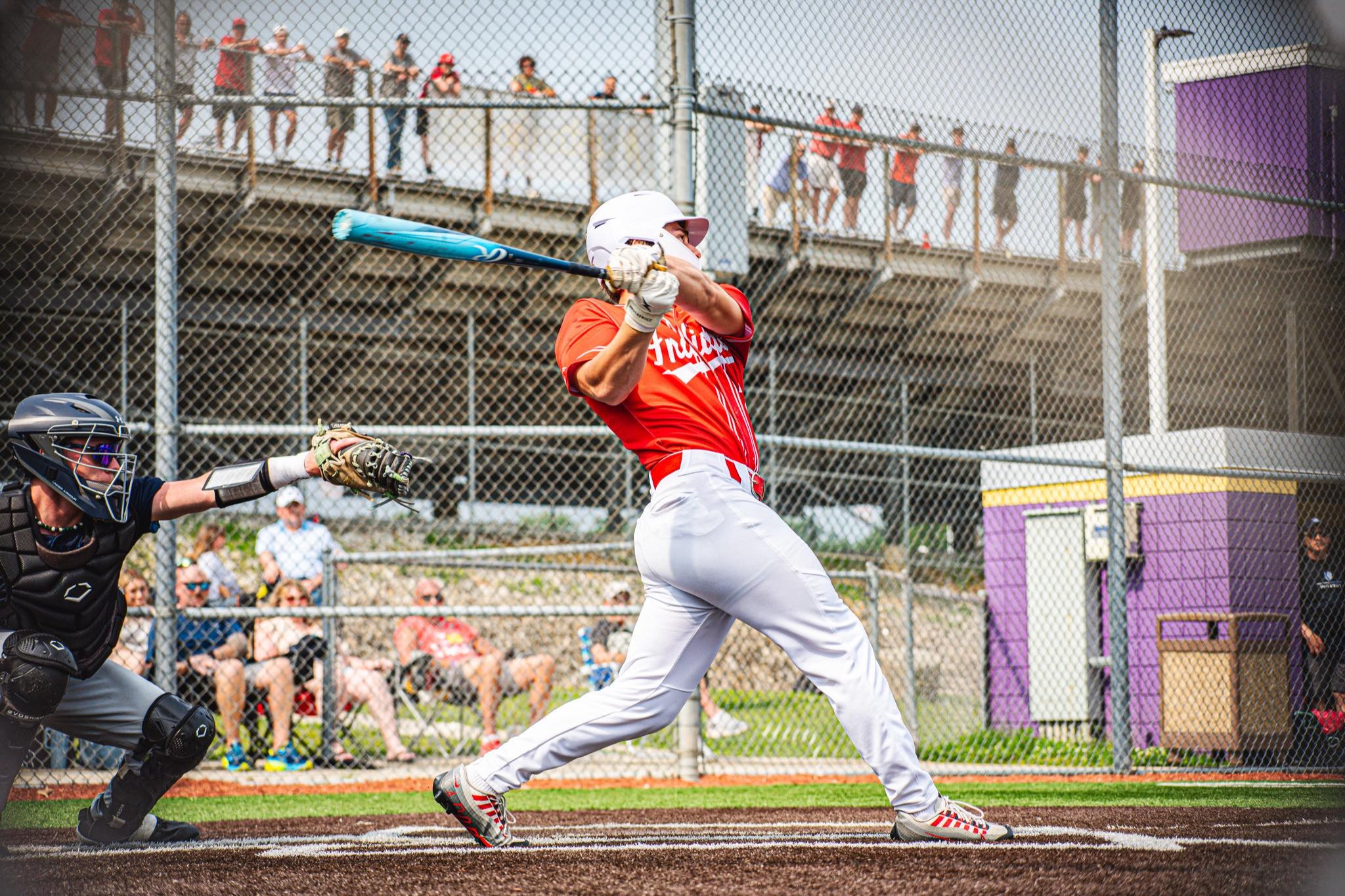 A baseball player swings a bat in an outdoor stadium. Spectators watch from behind a fence, and the umpire kneels nearby.