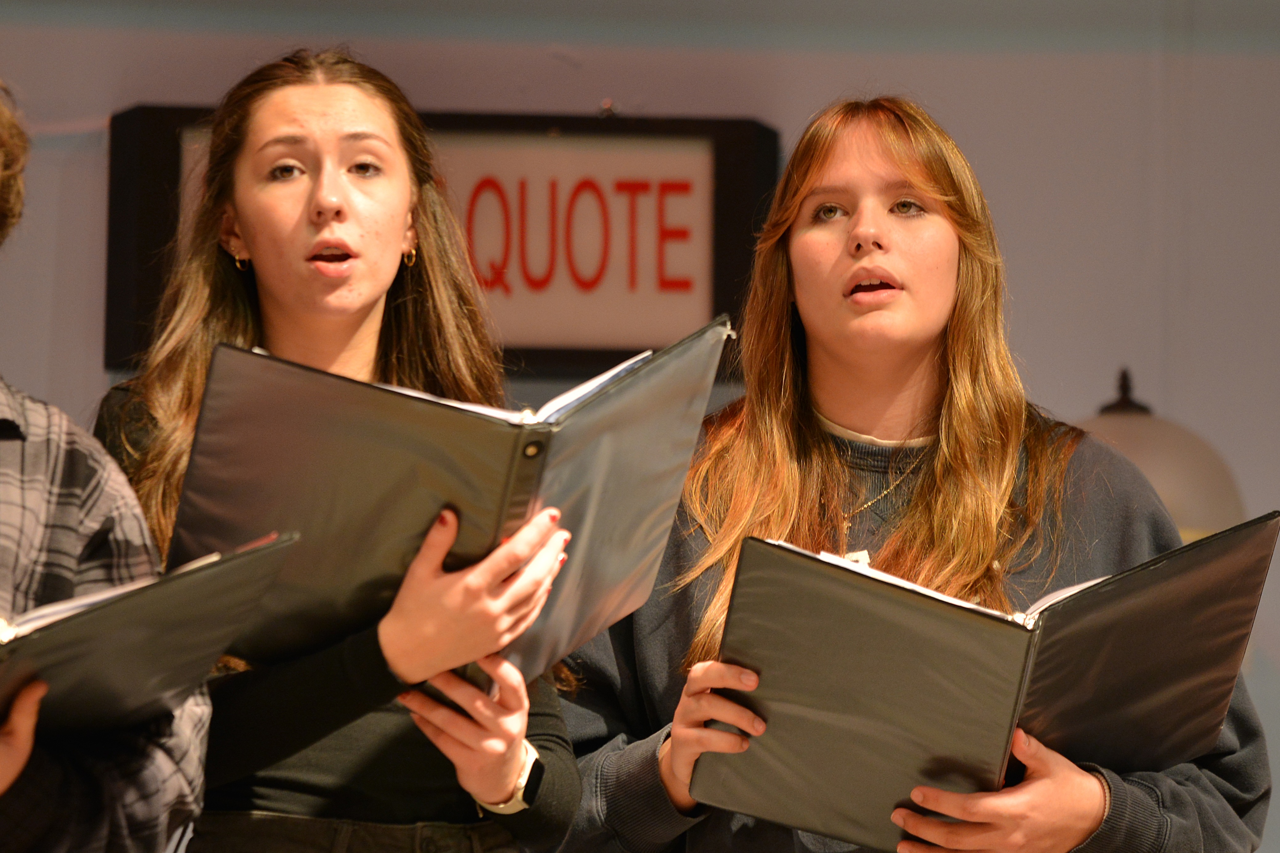 Three young adults in a classroom setting, holding binders and singing. A white sign with red letters reads "QUOTE".