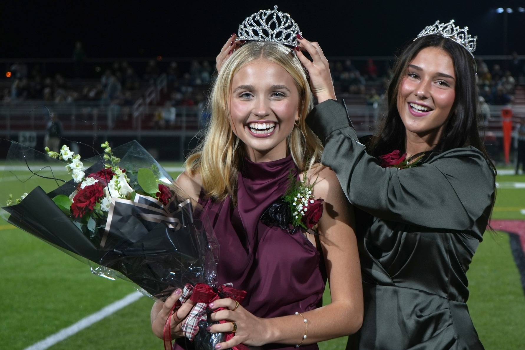 Two women on a football field, one wearing a crown and holding flowers, the other placing a crown on her head.