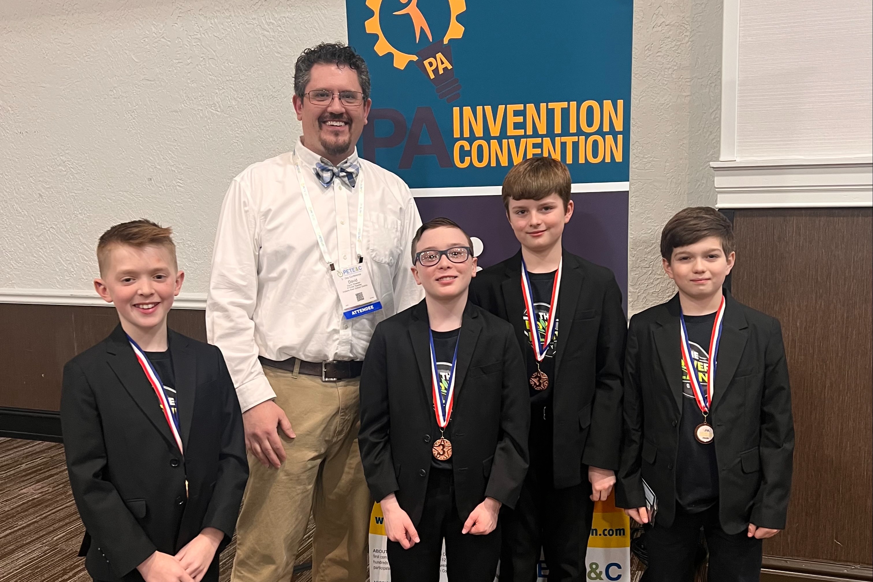 Five boys and a man wear medals. The man wears a bow tie and a white shirt. Behind them is a banner.