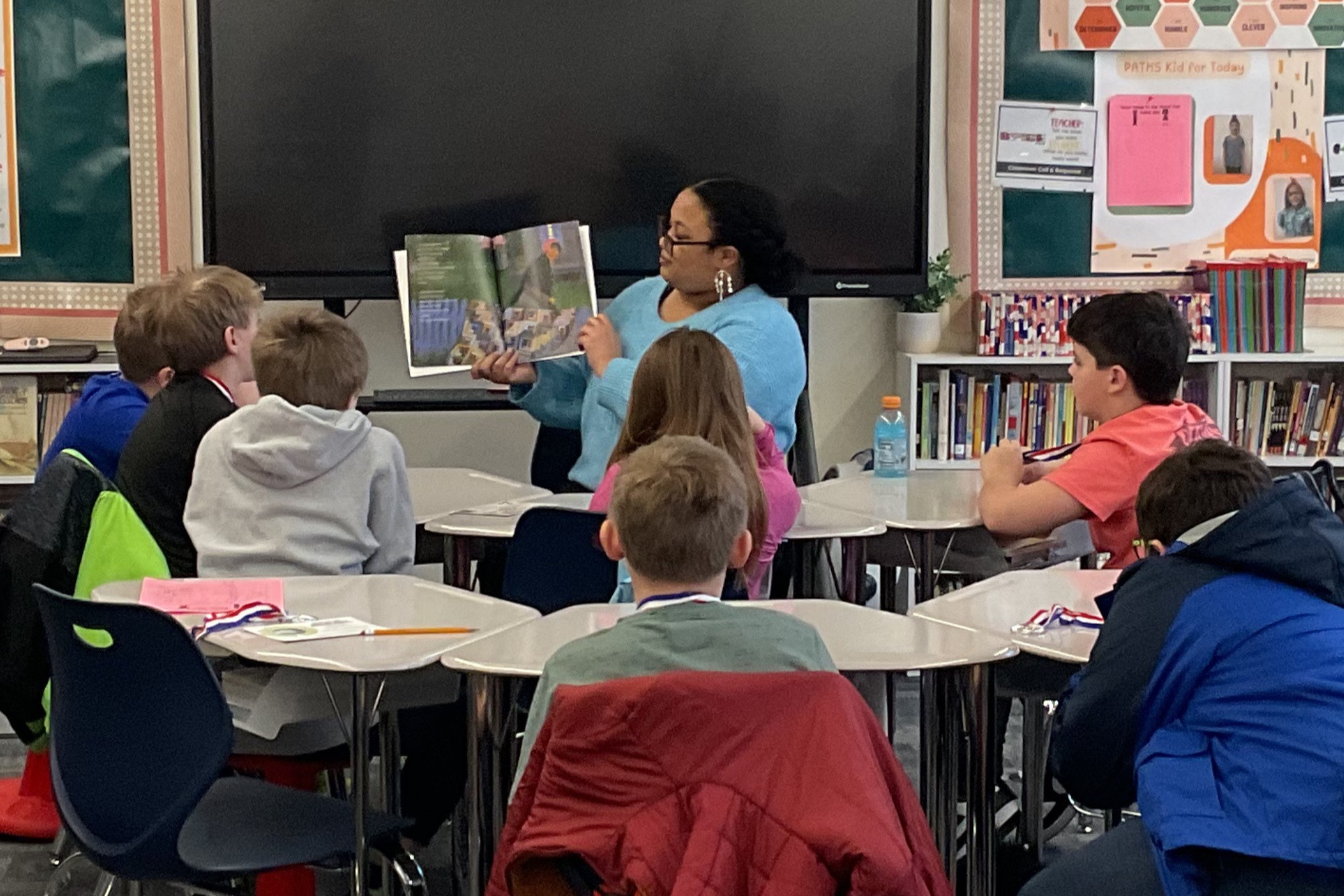 In a classroom, a woman reads a book to a group of students sitting around tables.