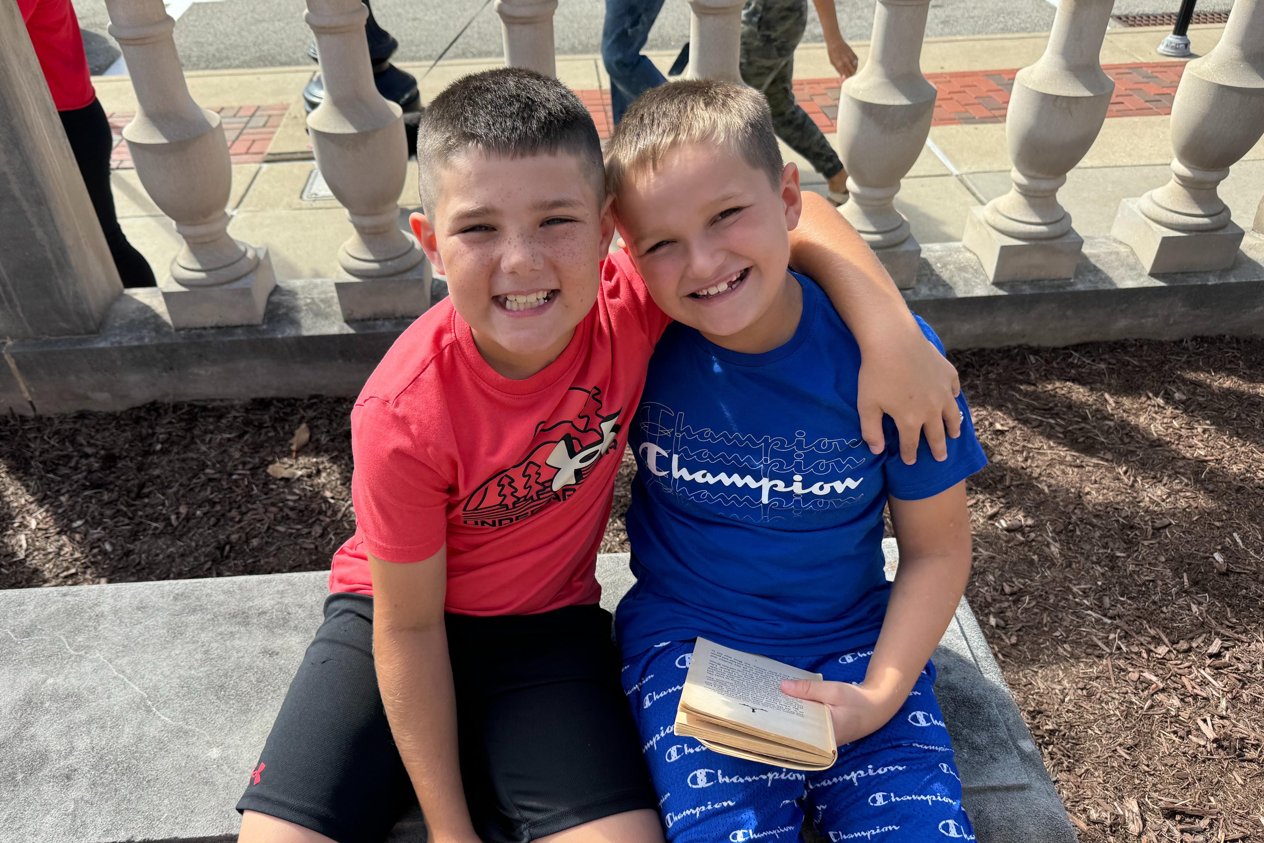 Two boys sit on a stone bench, smiling. One holds a book, the other holds a book. Behind them, a decorative fence with balustrades.