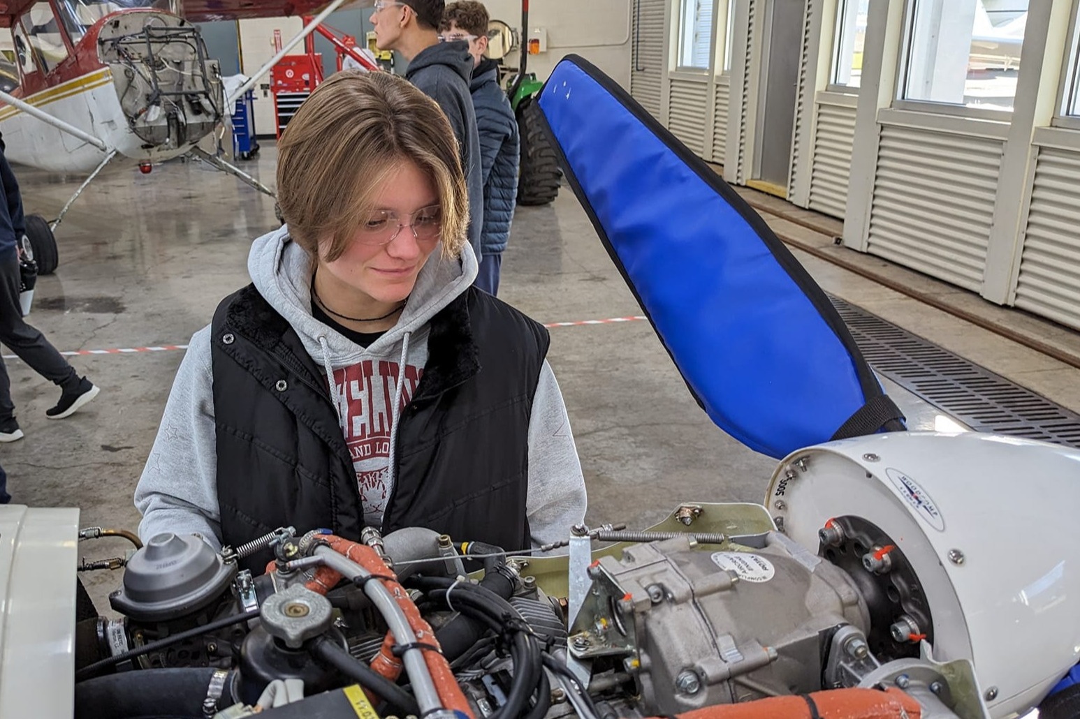 A person stands by a plane's engine, wearing a jacket. Other people are in the background.