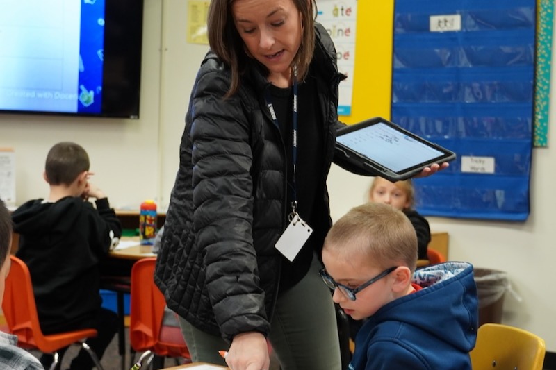 a teacher holding a tablet in a classroom setting helping a student