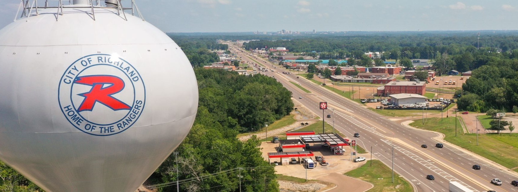 An aerial view of the City of Richland features the city's iconic water tower displaying the City of Richland logo and the words "Home of the Rangers." A major roadway lined with businesses stretches through the city, with green spaces, trees, and commercial areas visible under a clear sky.