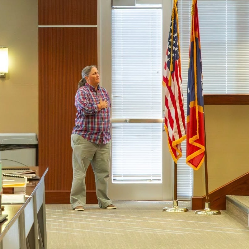 A person stands with their hand over their heart during the Pledge of Allegiance inside the Richland City Hall council chambers. The American and Mississippi state flags stand beside the speaker's platform, with meeting tables and seating visible in the room.