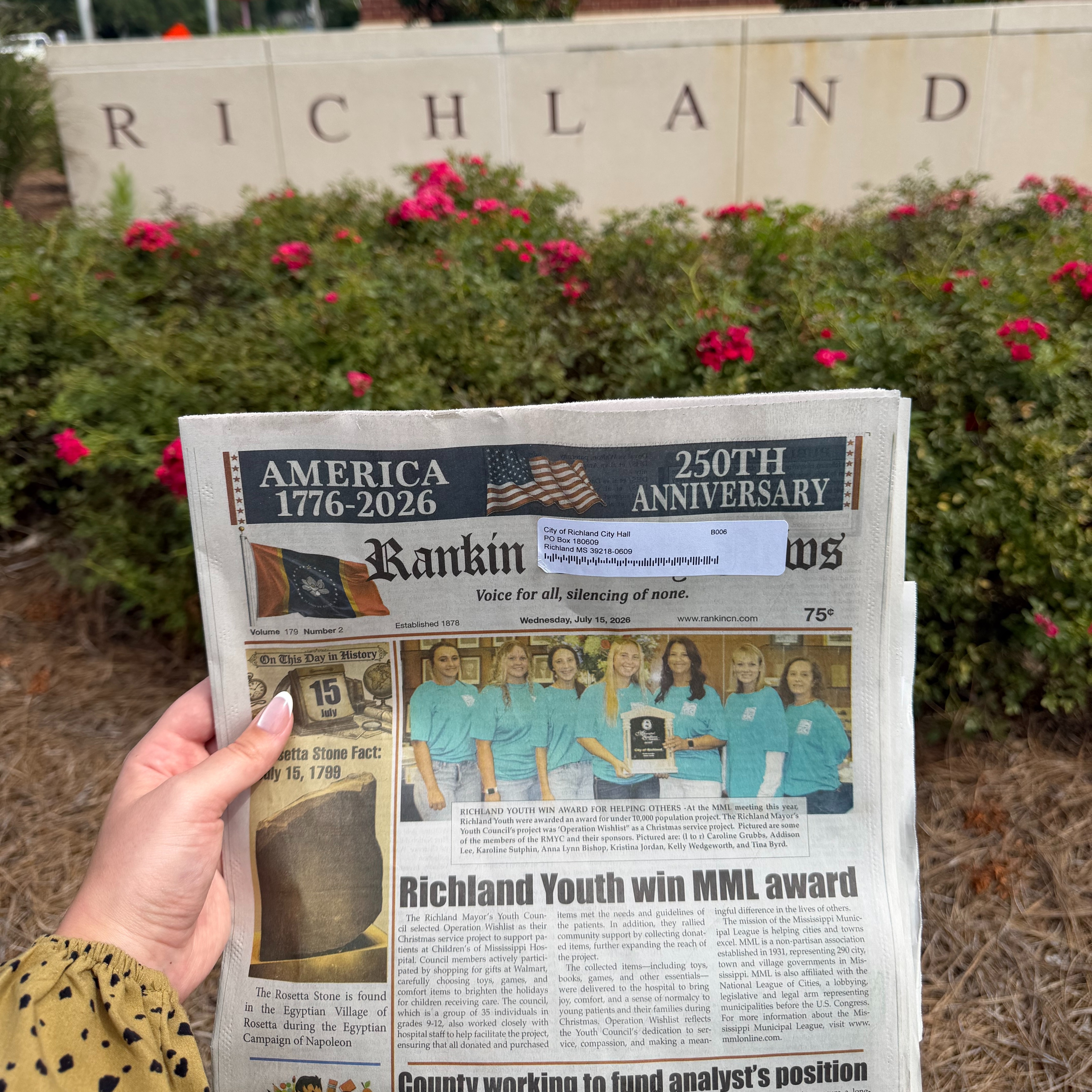A person holds a copy of the Rankin County News in front of the City of Richland entrance sign and landscaped flower beds. The newspaper's front-page headline reads, "Richland Youth win MML award," highlighting the Richland Mayor's Youth Council's recognition for its community service project. City Hall and flags are visible in the background
