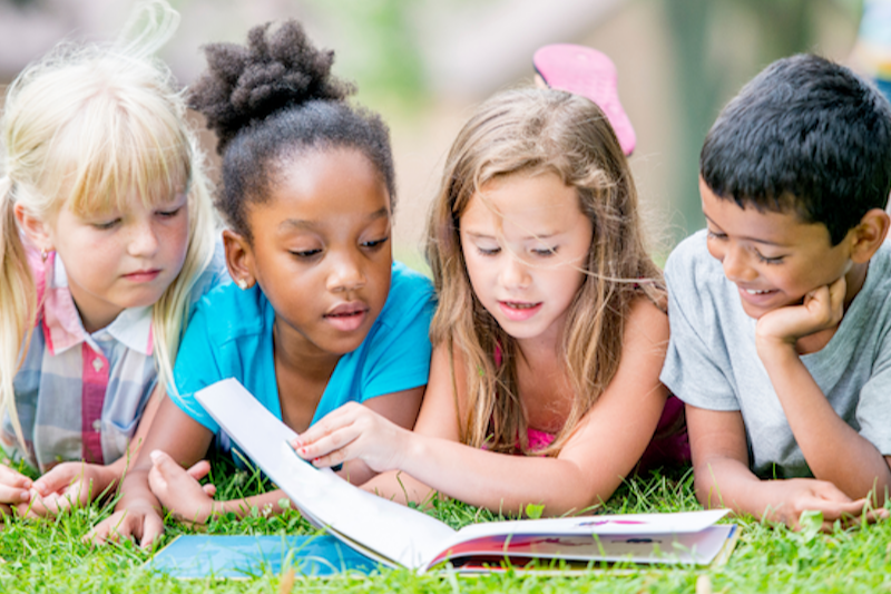 four school aged student lying in the grass all reading the same book