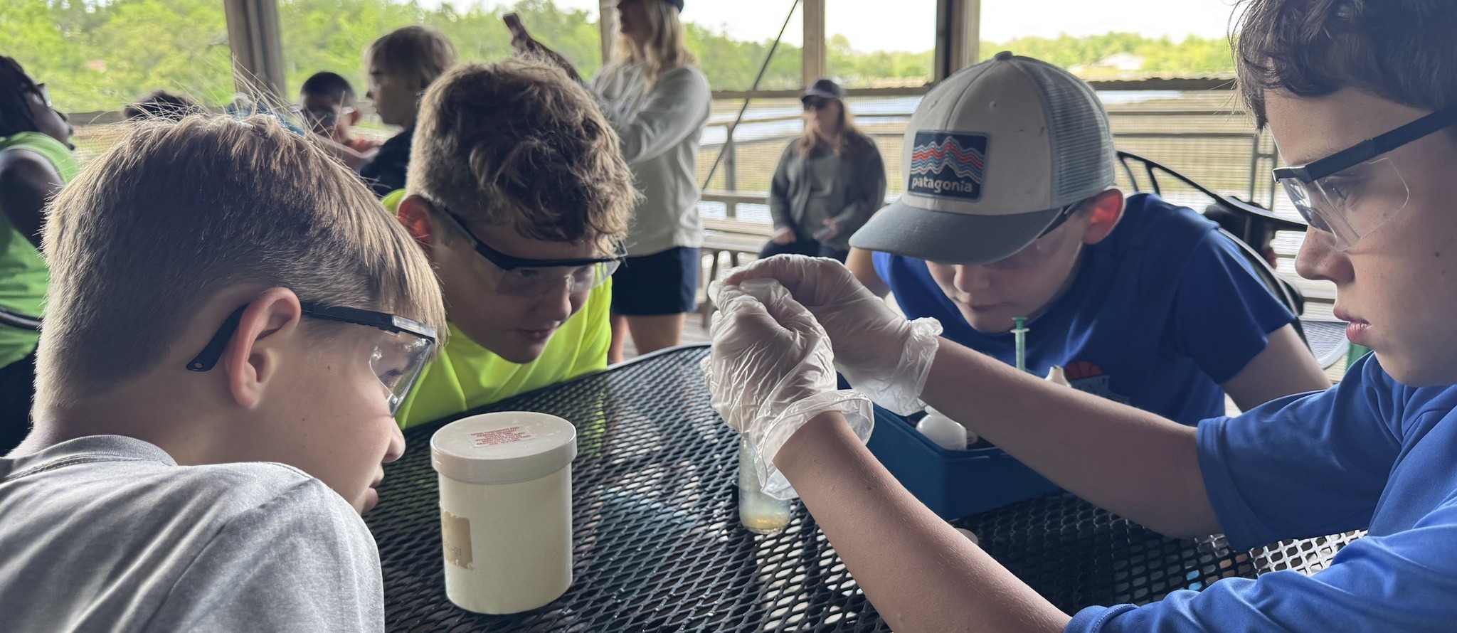 Students sitting at a table leaning in working on a science project outside with safety glasses