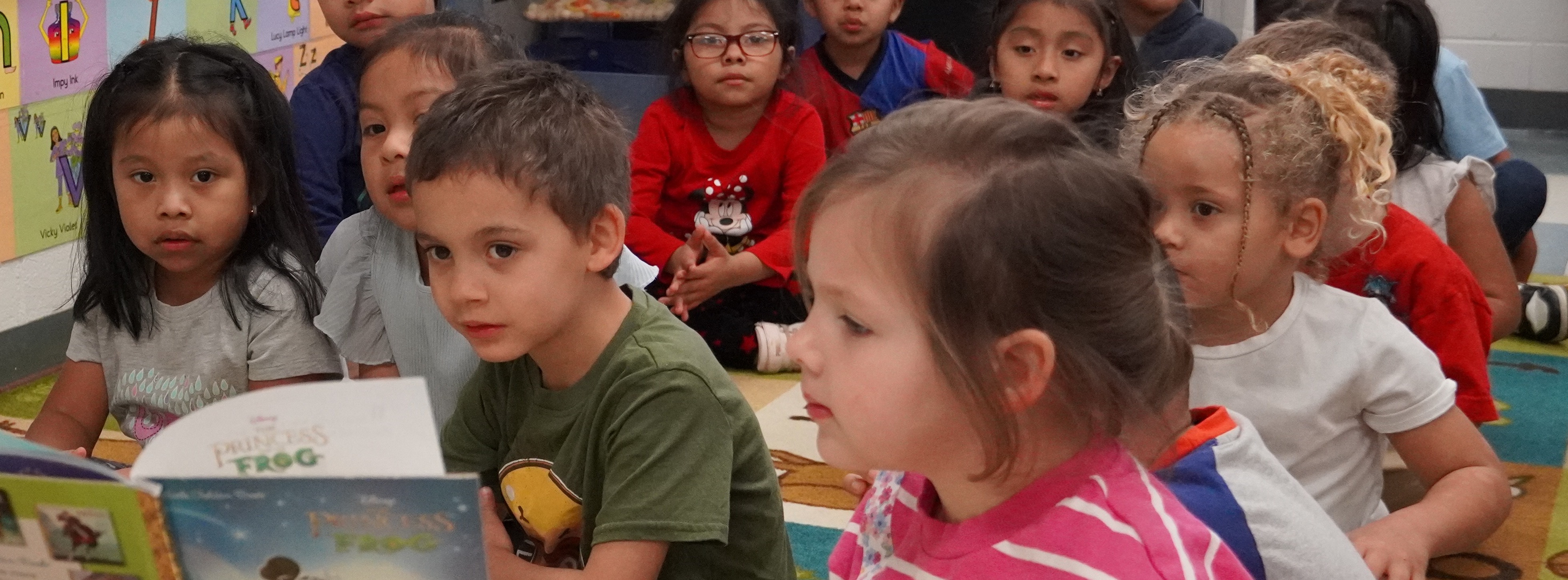 adult reading a book to a group of students sitting on the floor of a classroom