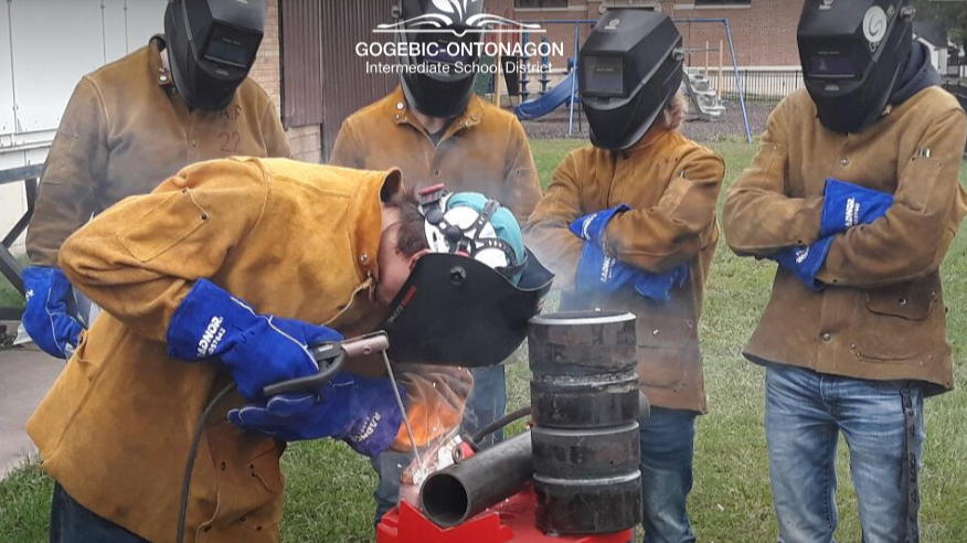 High school students observing a classmate demonstrating a welding technique outdoors. All are wearing welding helmets and protective leather jackets and gloves.