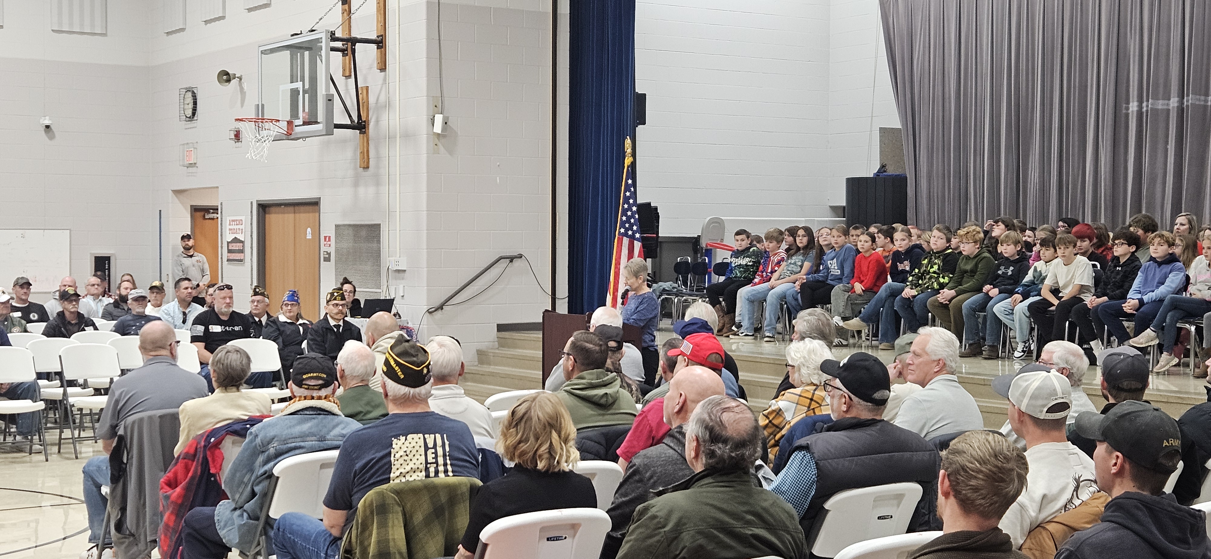 Large group of people sitting in chairs in a hall with a stage and a flag.
