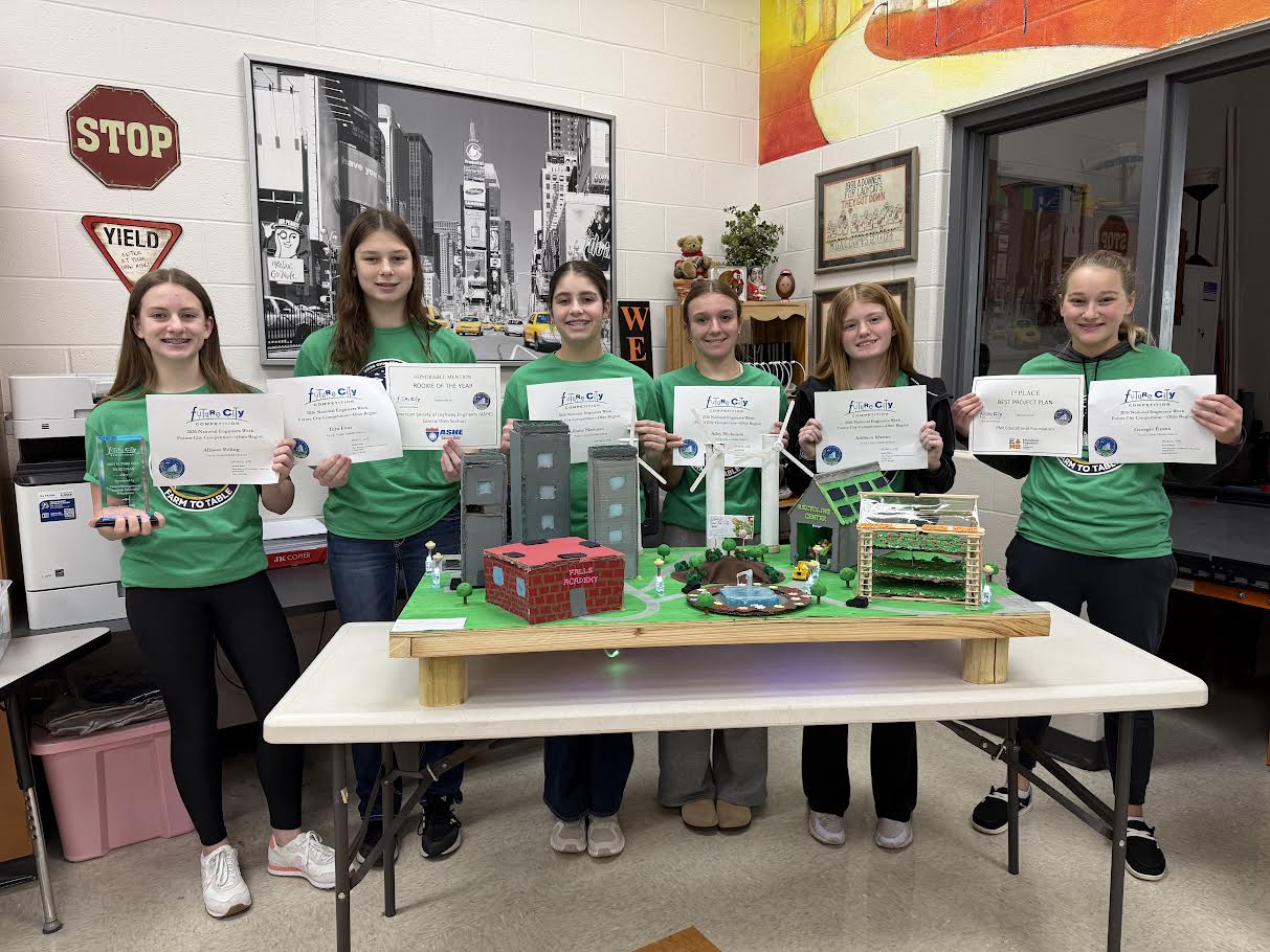 Six young girls, in matching green shirts, pose with certificates. Behind them, a table holds a miniature landscape model.
