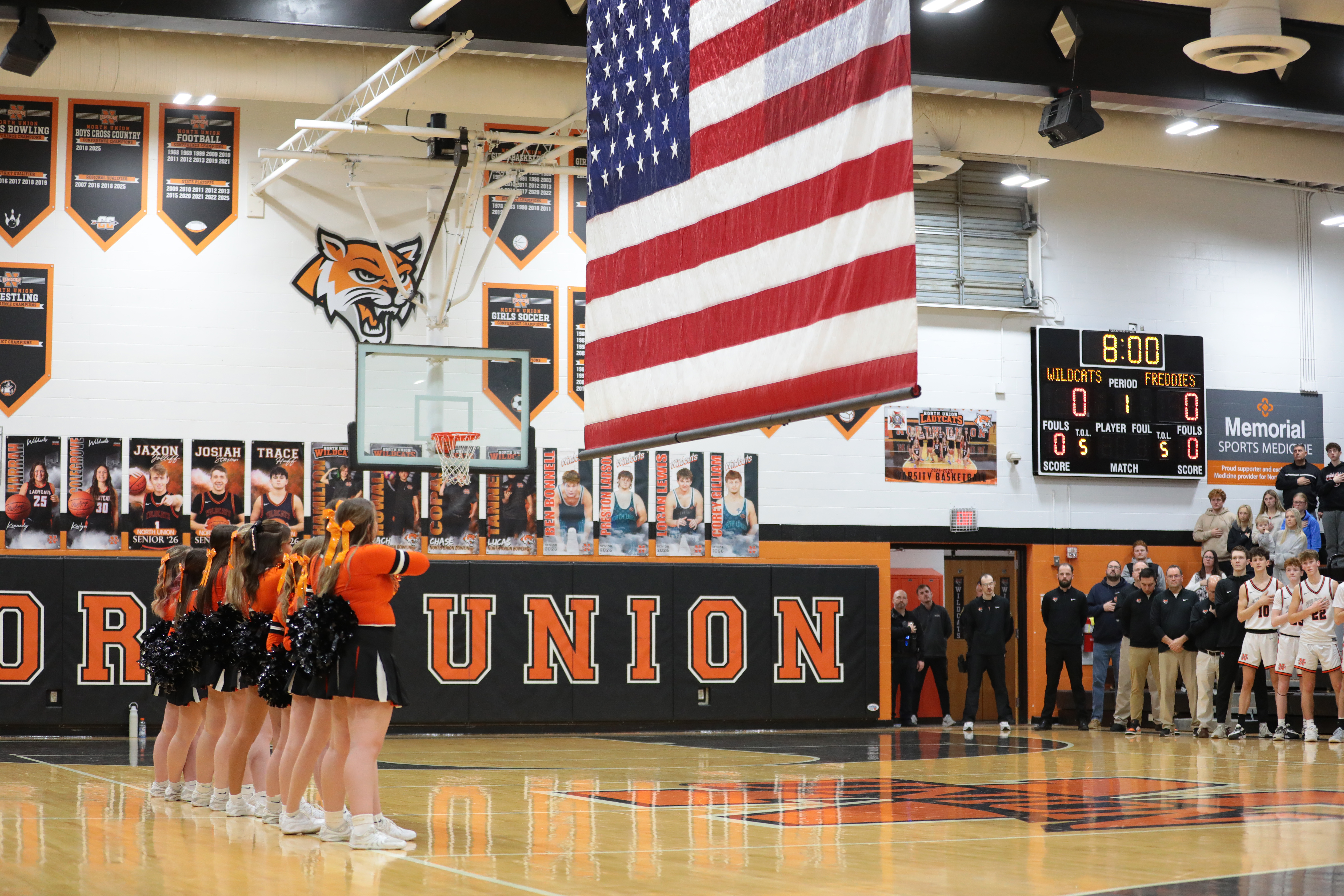 A basketball court with cheerleaders lined up. An American flag hangs overhead. The wall reads "DR UNION".