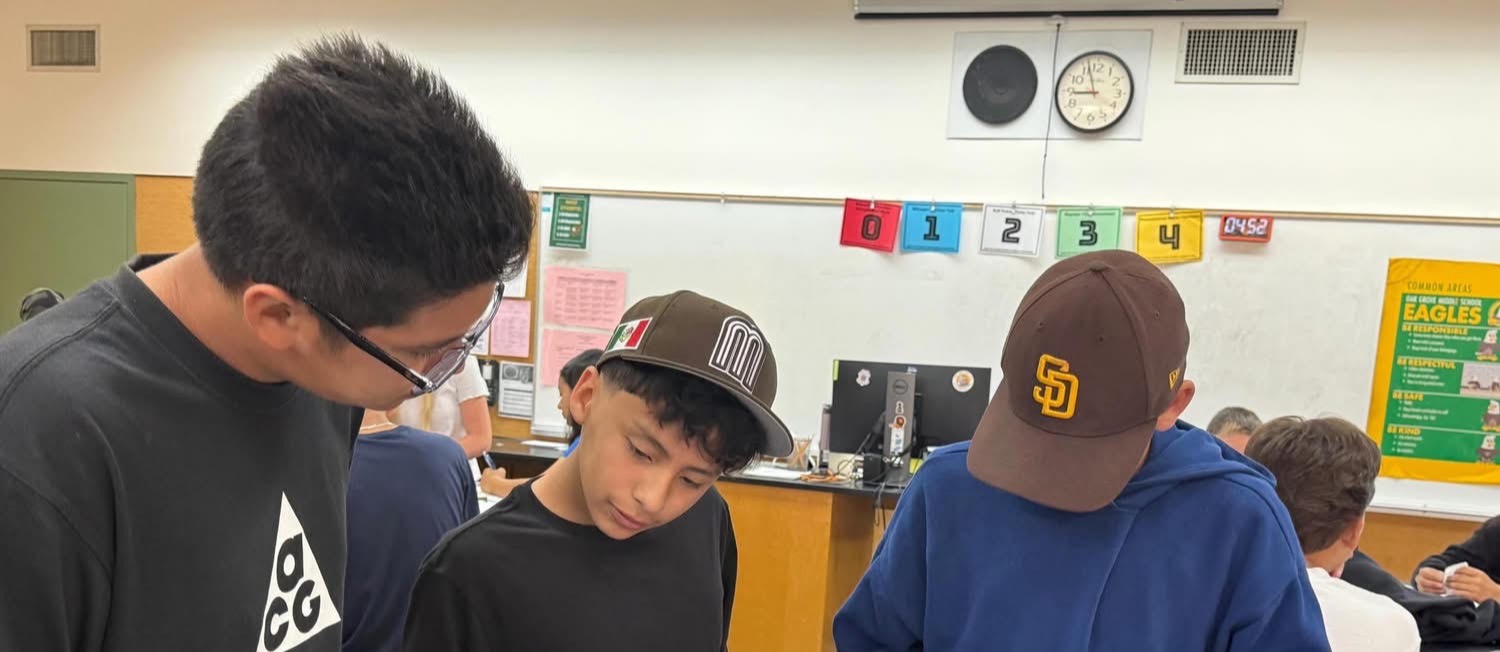 Three students work on a table with papers and pencils; a classroom background has a whiteboard and bulletin board.