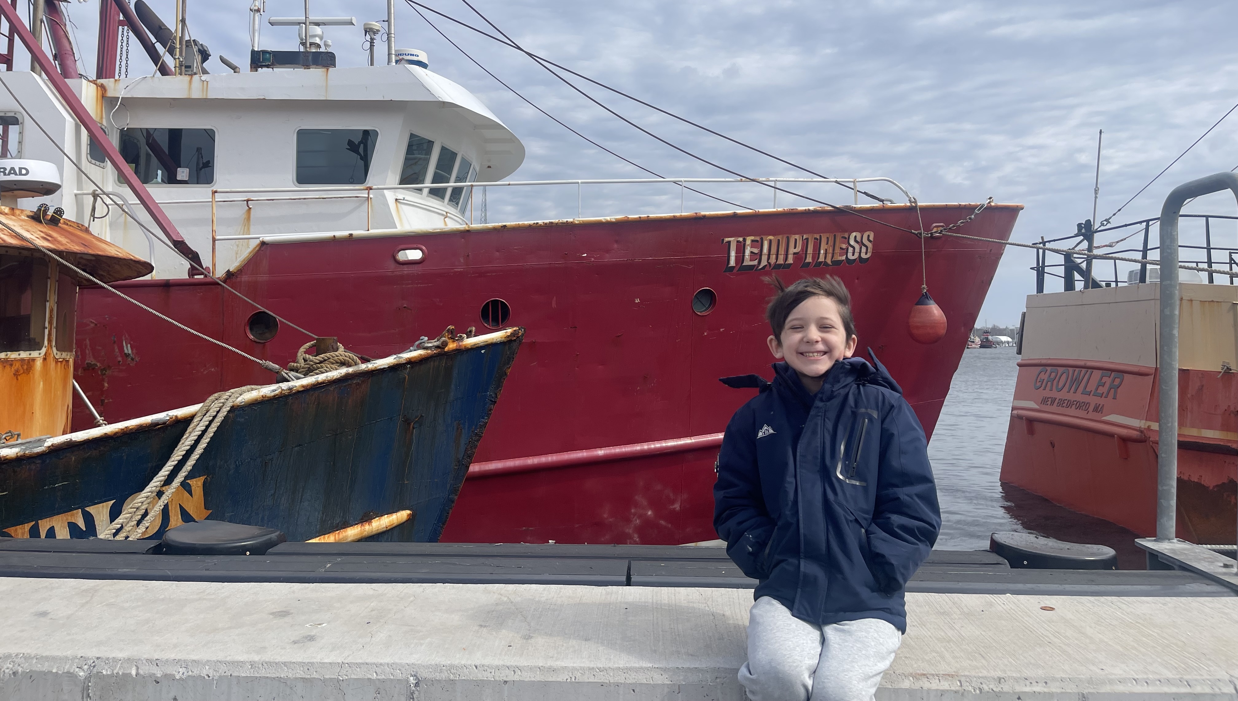 a smiling child at a pier standing in front of a fishing boat