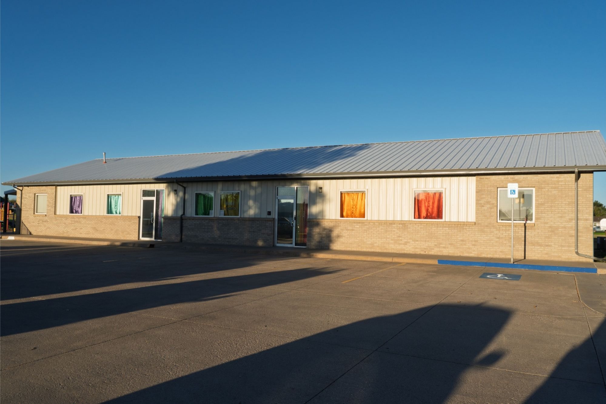 A building with a metal roof has a parking lot in front. Large windows display colorful artwork.