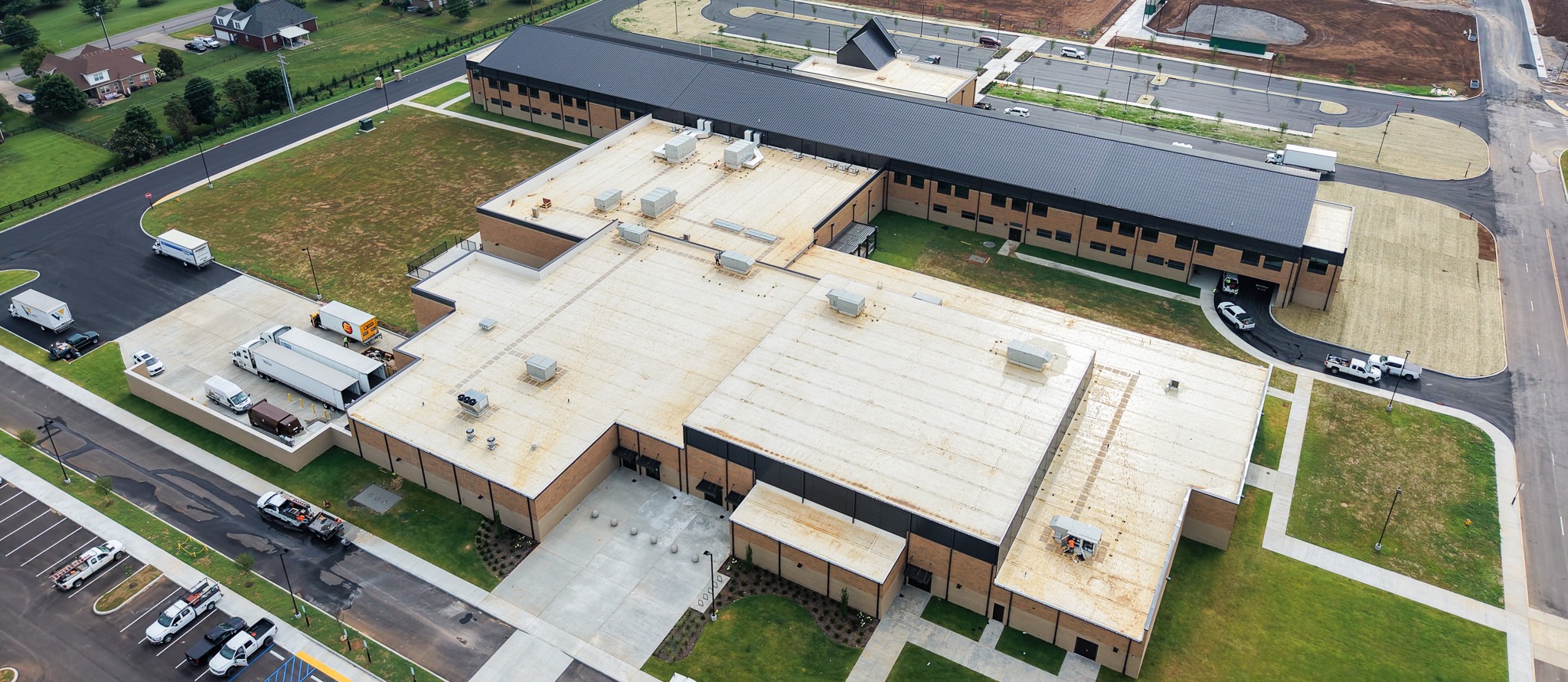 Aerial view of a large building with a parking lot. Surrounding area includes fields, houses, and a road.