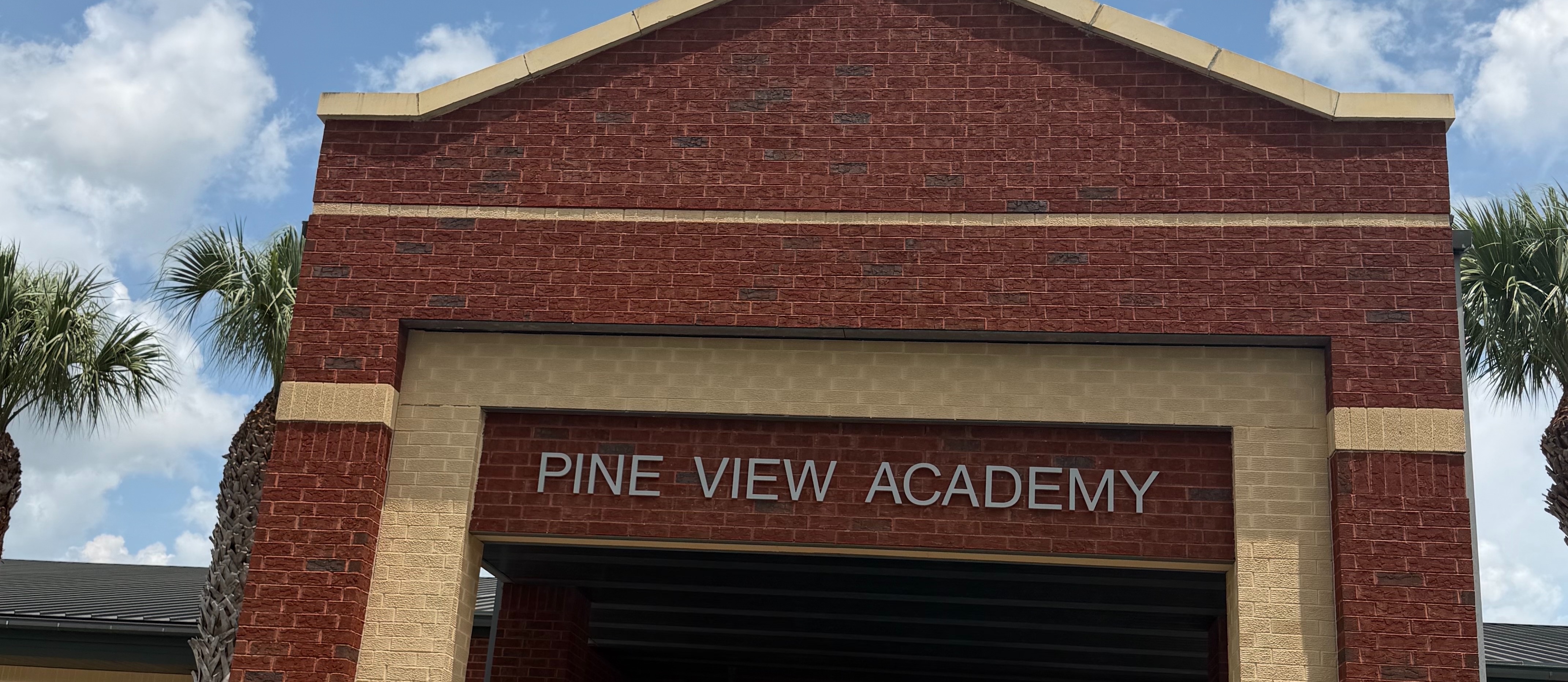Entrance to Pine View Academy, a red brick building with arched doorway. Green lawn and palm trees in the background.
