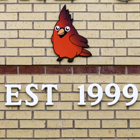 Red cardinal with "Est. 1999" written below in white letters against a brick wall. 