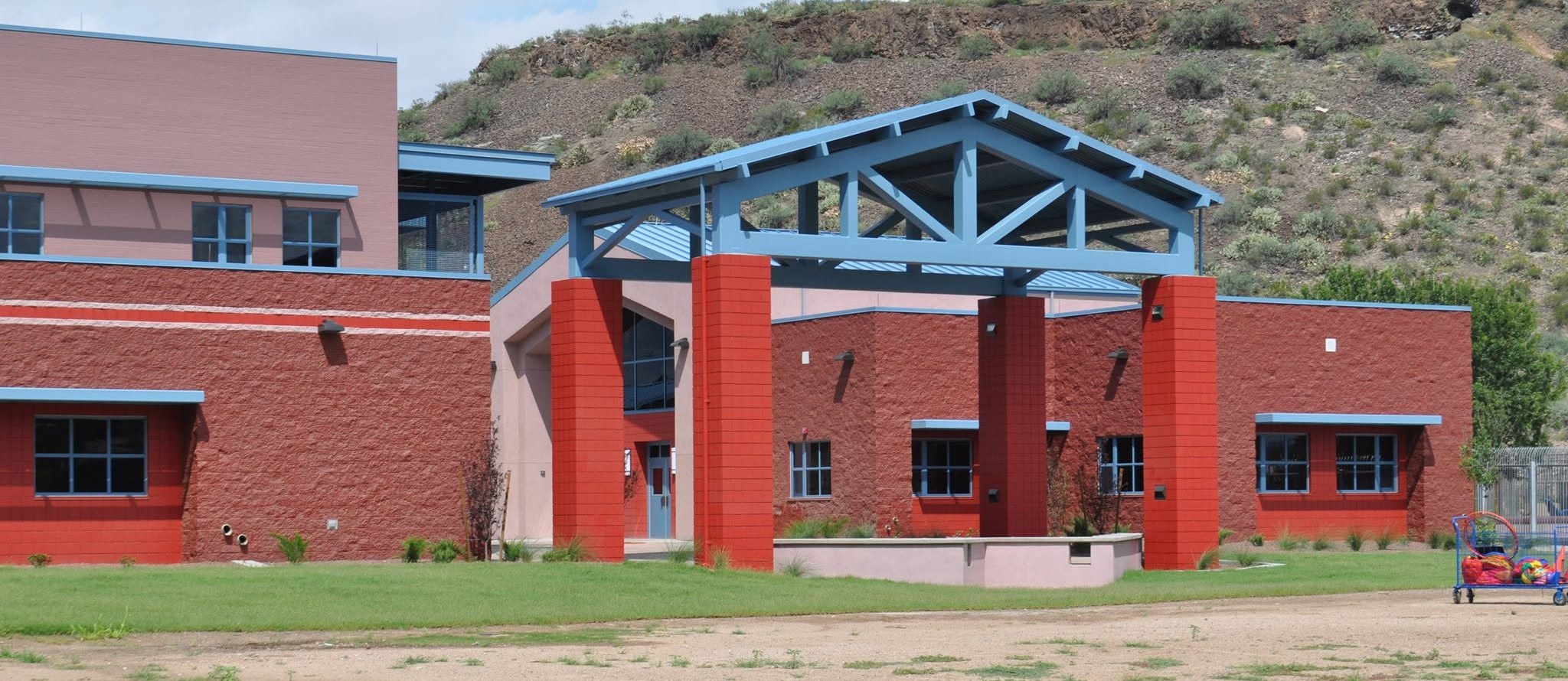 Red building with a blue roof and red pillars in front. Grassy area with dirt patches. Mountain backdrop.