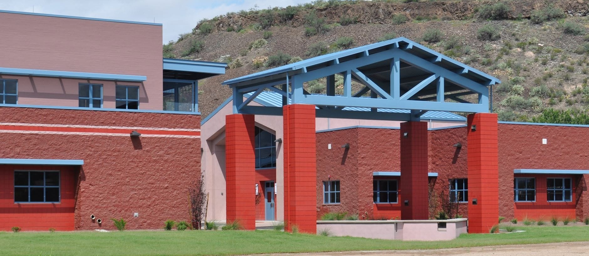 Red building with a blue roof and red pillars in front. Grassy area with dirt patches. Mountain backdrop.