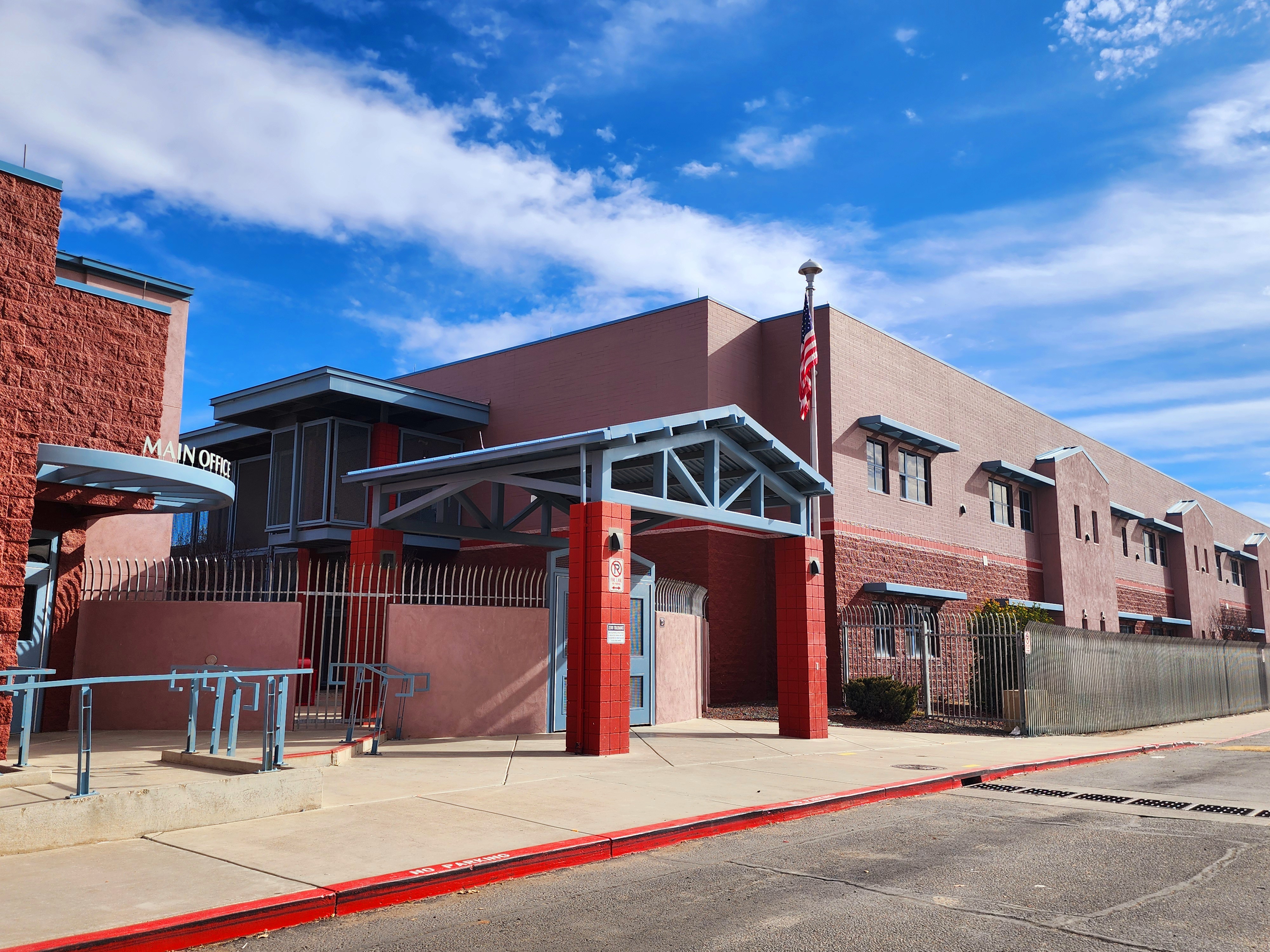 A large building with a red facade, a flag, and a metal canopy stands on a street with a red curb.