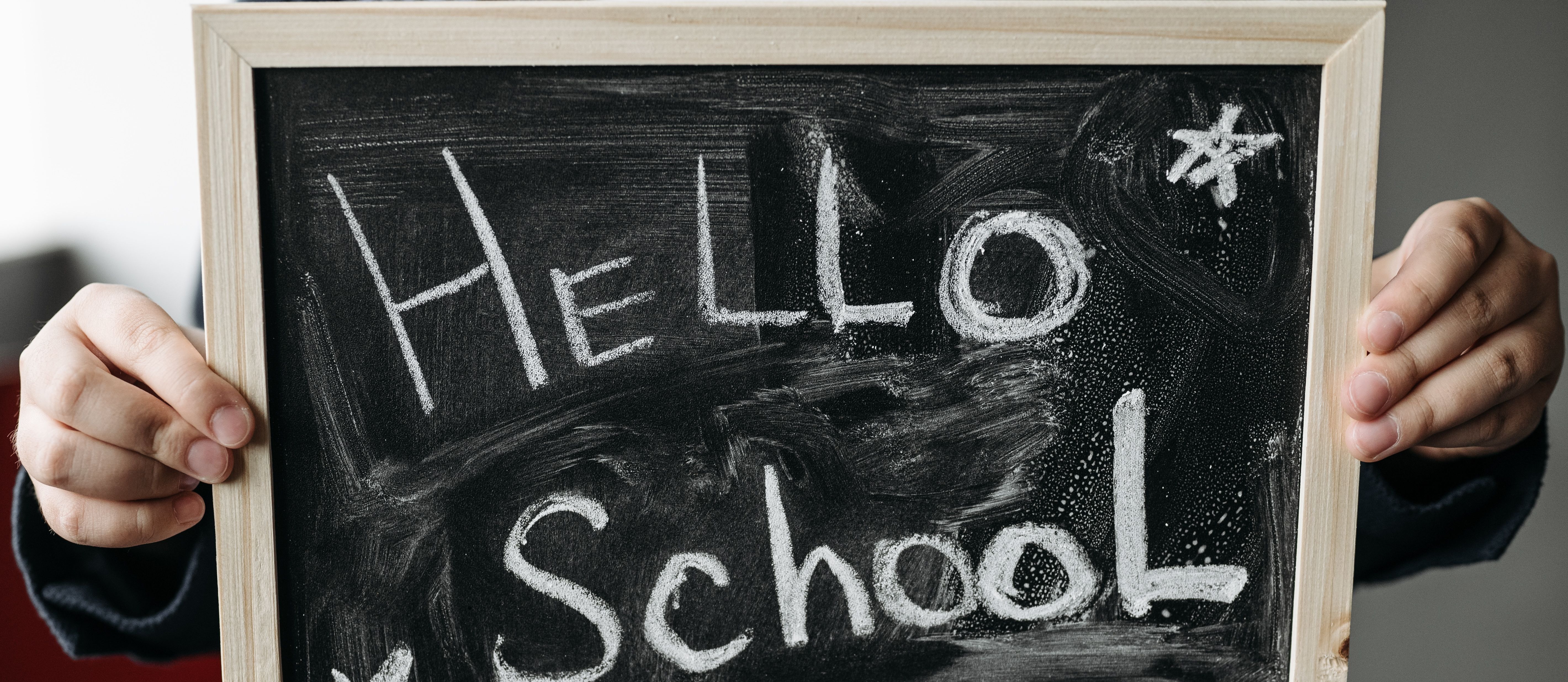 A person holding a framed chalkboard with "HELLO School" written in white chalk. Red background.