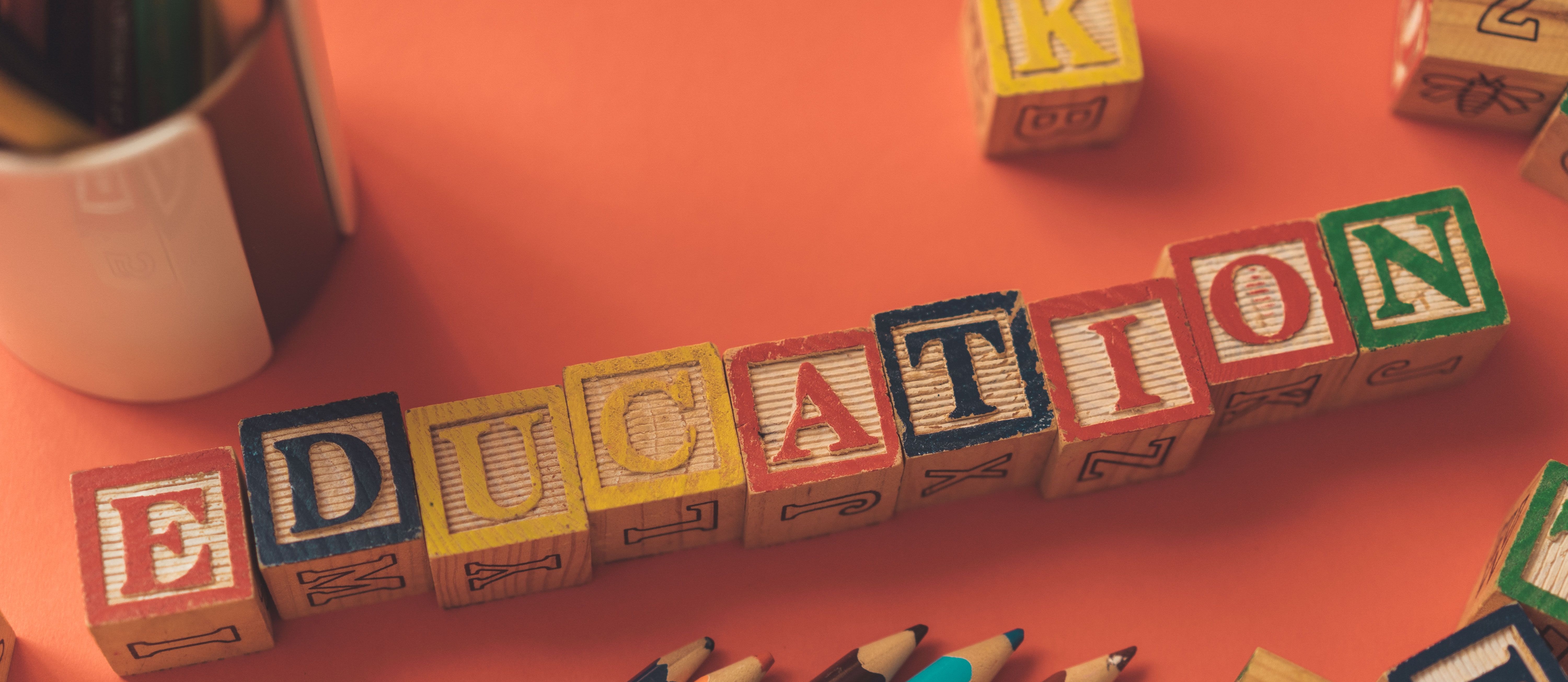 Orange background with wooden blocks spelling "Education" and assorted colored pencils. A pencil holder is visible.