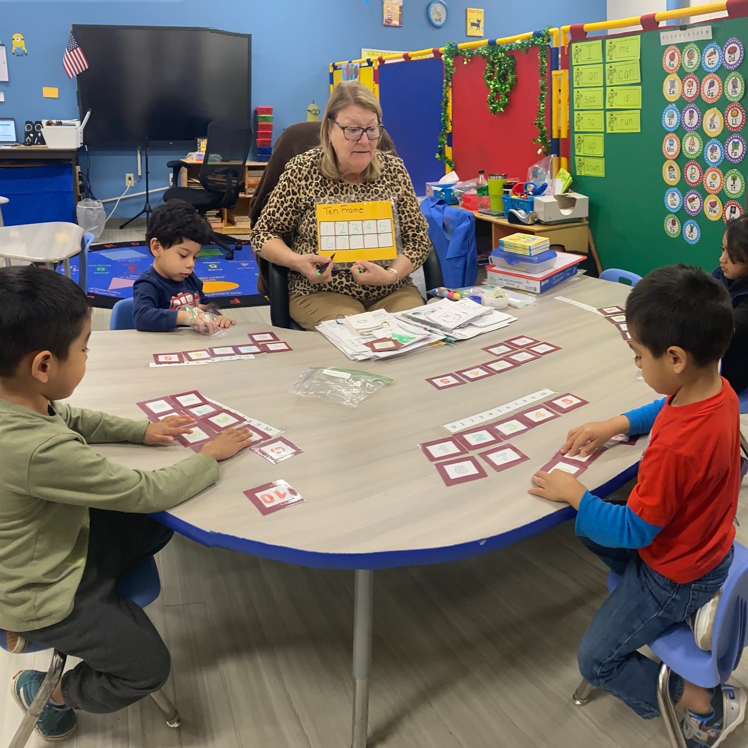 Teacher and four children engage in a card activity around a round table in a colorful classroom.